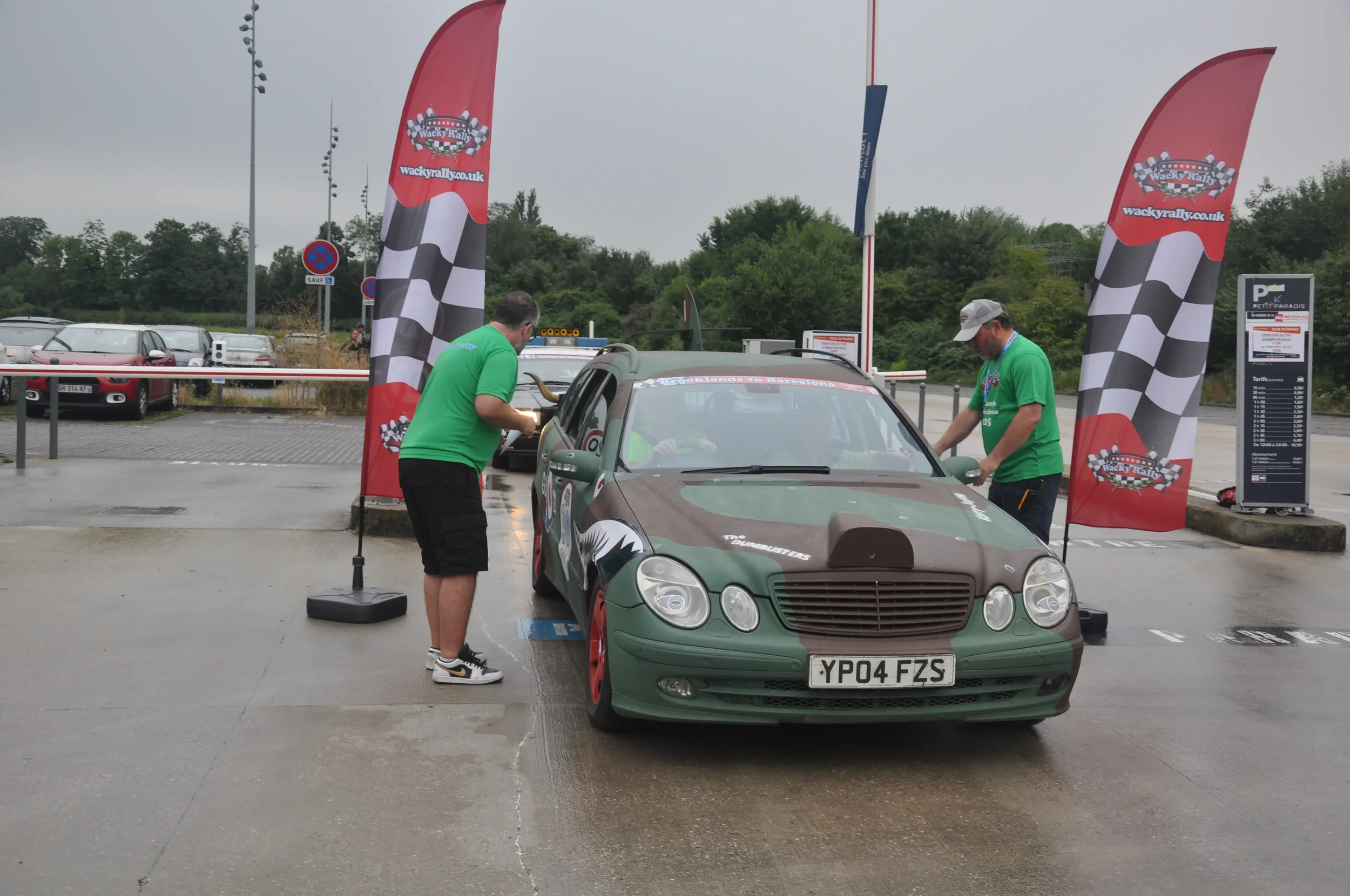 A rally car with a camouflage paint job is parked in a designated area, with two crew members in green shirts working on the car. The area is marked with red banners and checkered flags, and the scene appears to be at a car circuit or rally event in 