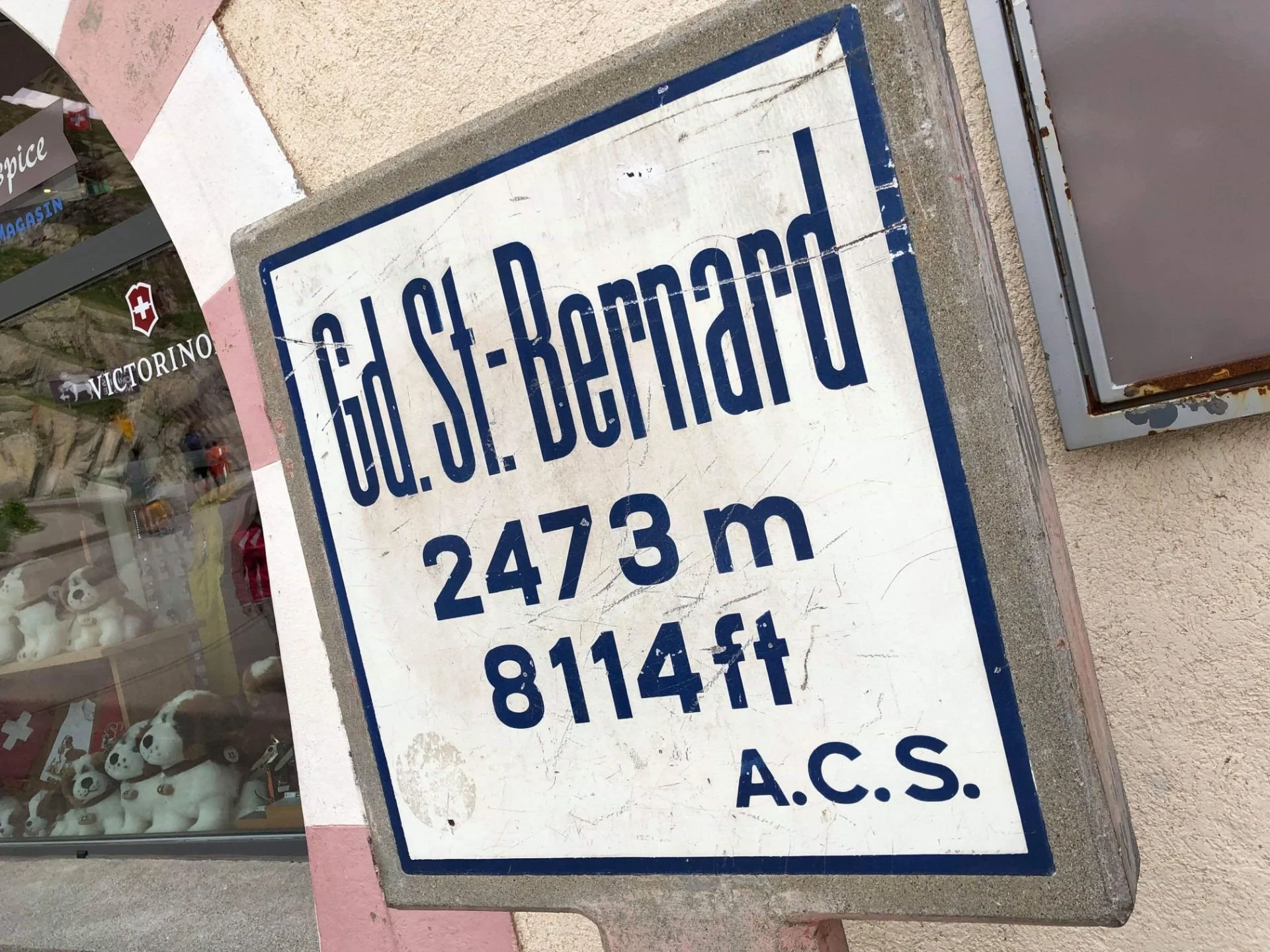 Street sign indicating the location of the Gd. S. J. Bermudo at 2473 meters or 8114 feet elevation, mounted on a concrete wall outside a store with plush toys and a Swiss flag sticker