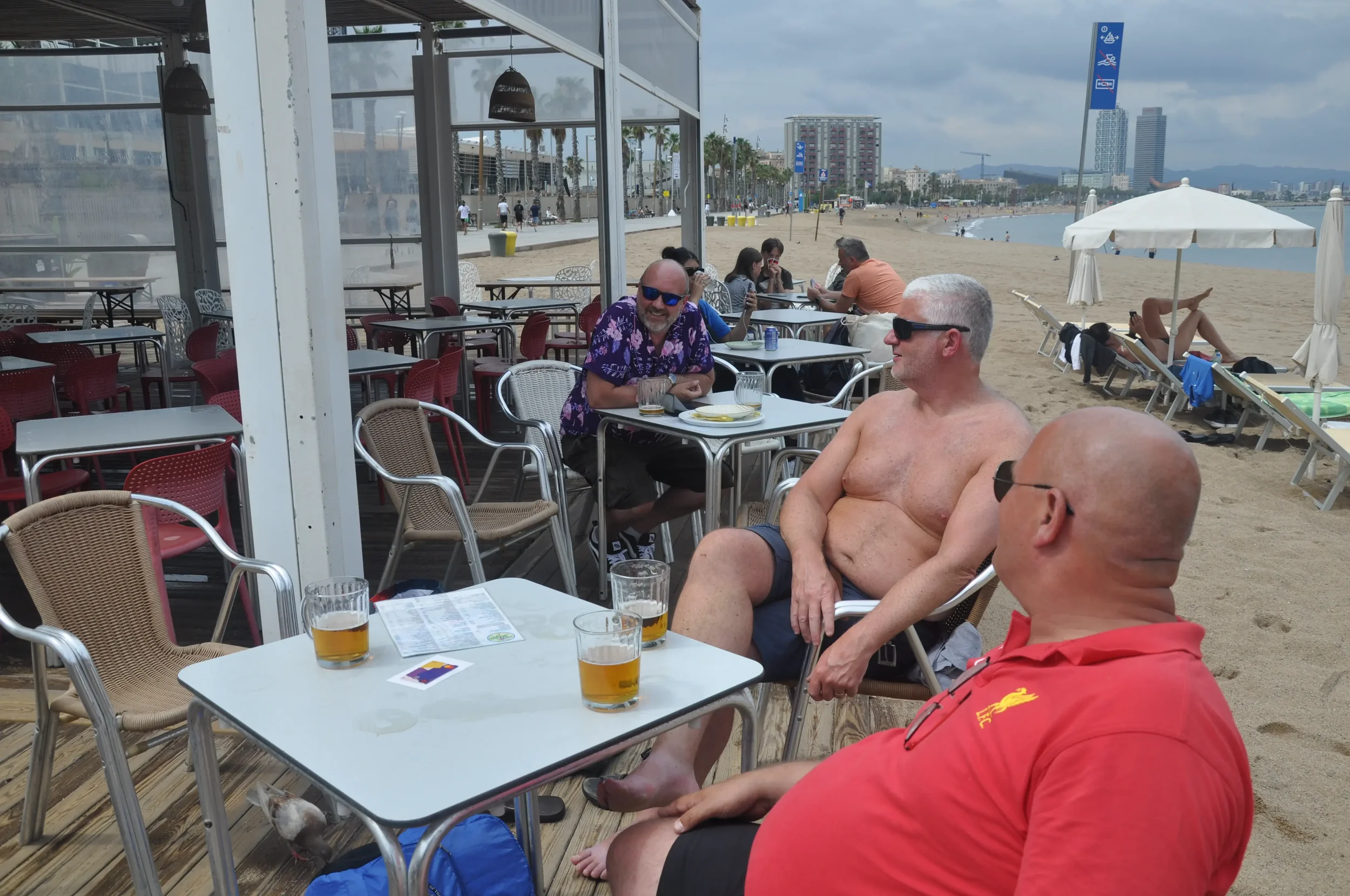 Three men sitting at a beachside restaurant, two wearing sunglasses and one shirtless, engaged in conversation with drinks on the table, with a beach and people relaxing in the background.