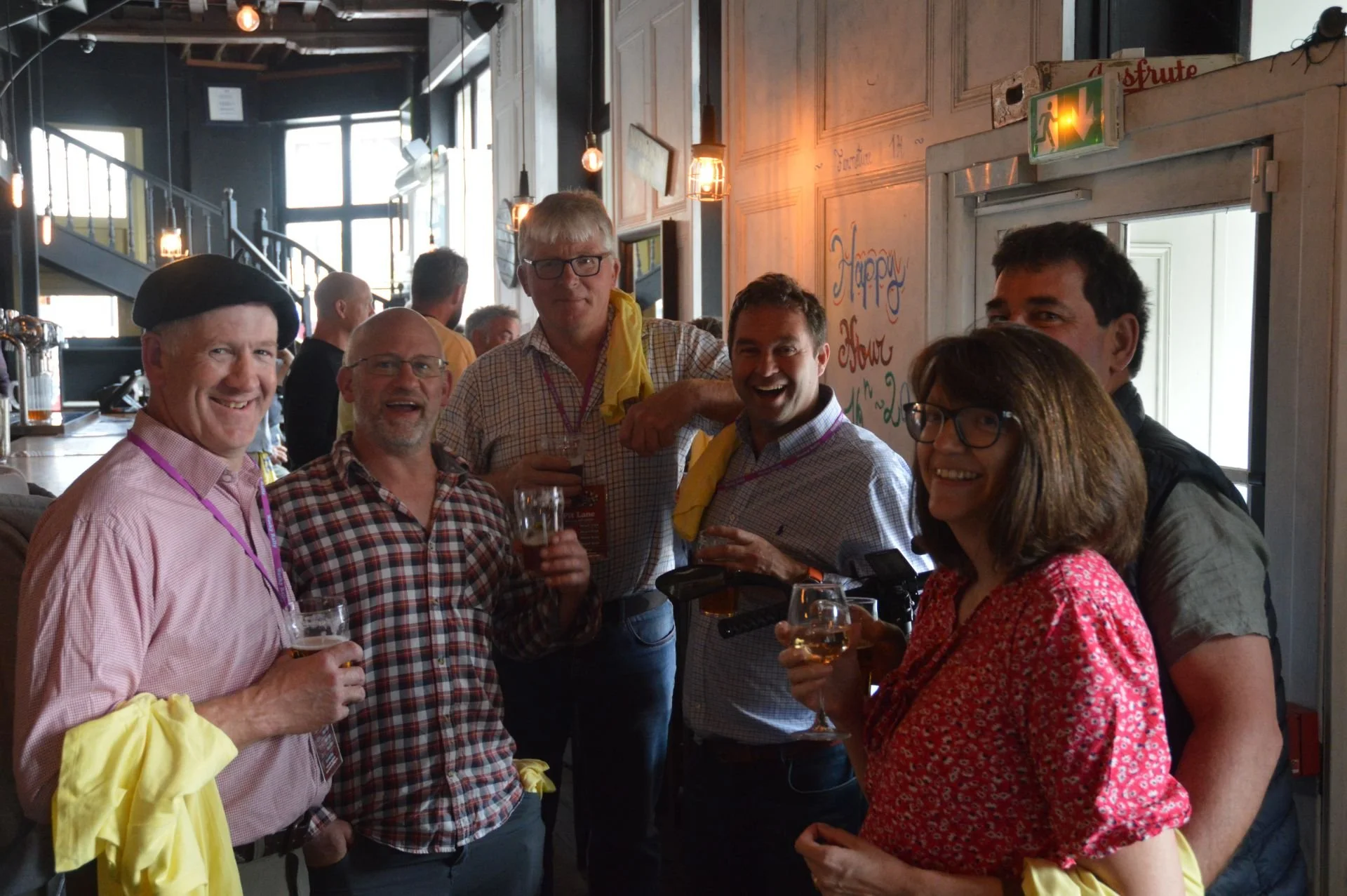 Group of six people celebrating at a bar or restaurant, holding drinks and smiling, with a festive sign on the wall behind them that says 'Happy Hour'.
