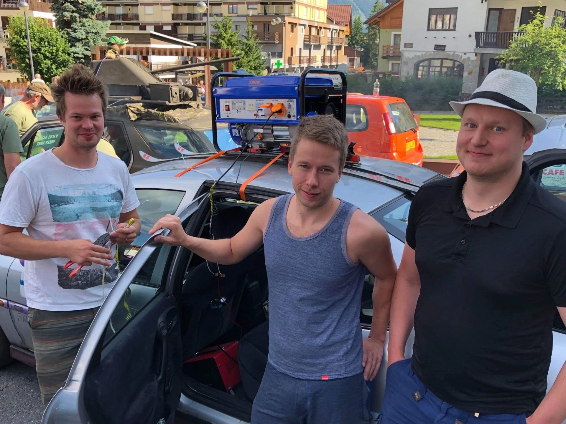 Three young men standing near a silver car, with one of them holding tools, in an outdoor setting with other vehicles and buildings in the background