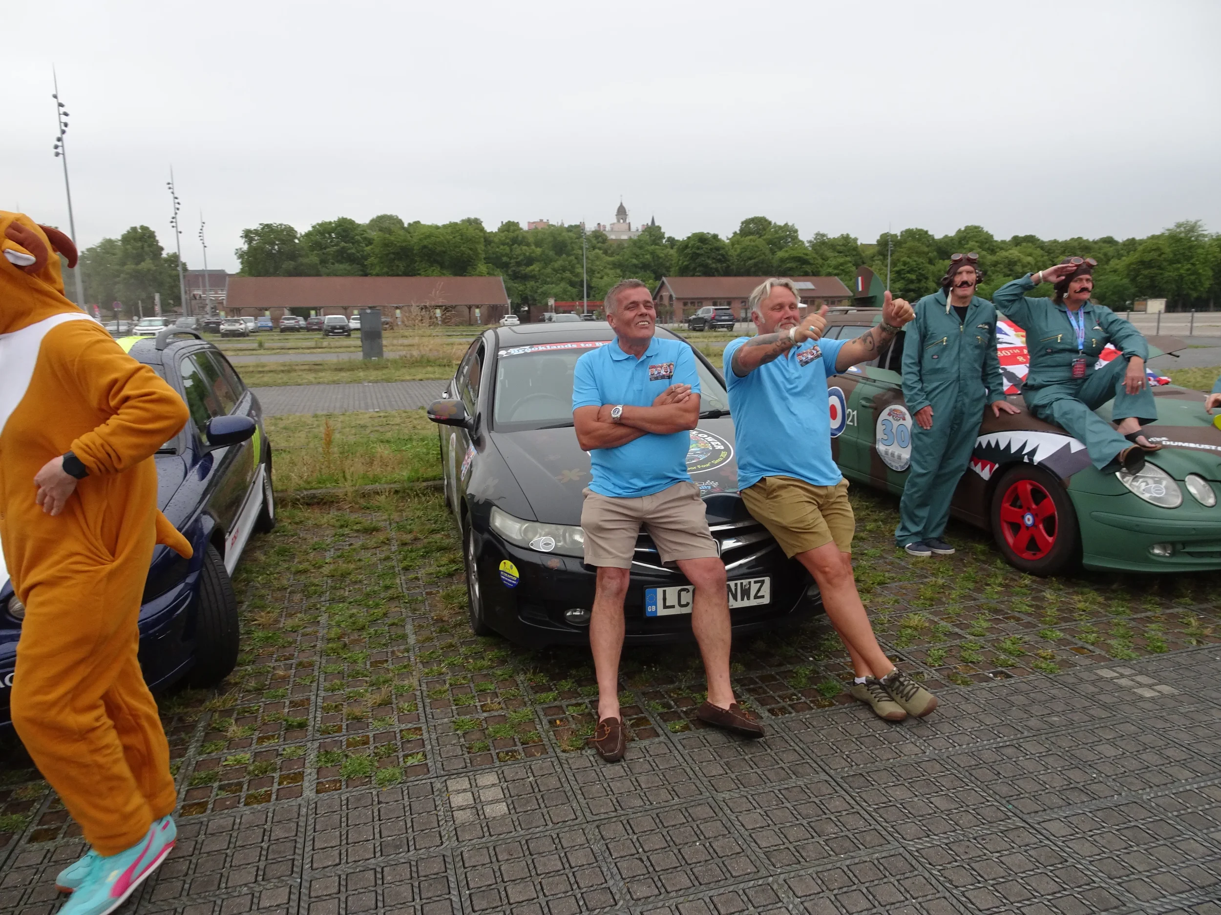 People standing and sitting near cars in a parking lot, with some dressed in costumes and casual clothes, under an overcast sky.