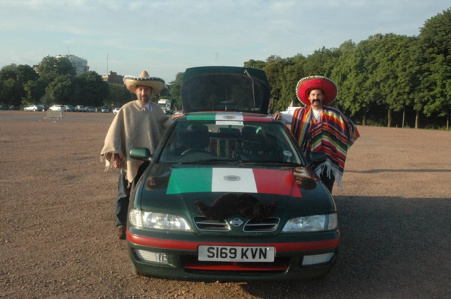 Two men dressed in traditional Mexican attire stand beside a black Nissan car decorated with the Mexican flag. One man wears a poncho and a sombrero, the other wears a colorful striped poncho and a red sombrero. The car's hood features a large bear a
