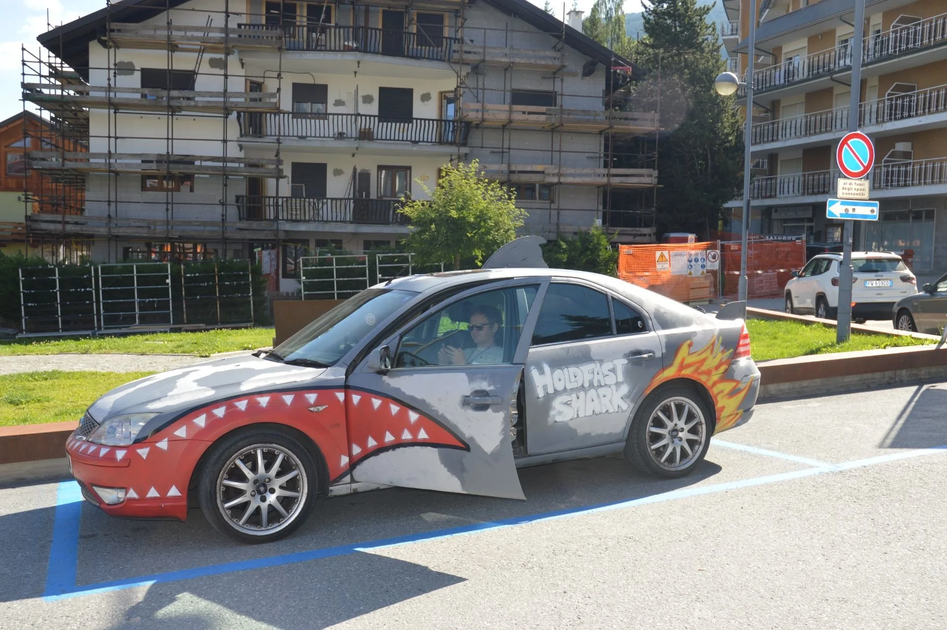 A parked car with a shark design on the side and the words "Hold Fast Shark" on the door, in a parking lot with buildings and trees in the background.