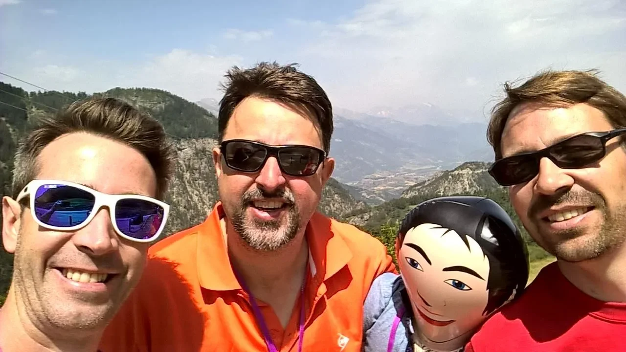 Four men smiling outdoors in a mountainous area, wearing sunglasses, with a cartoon doll and mountains in the background.
