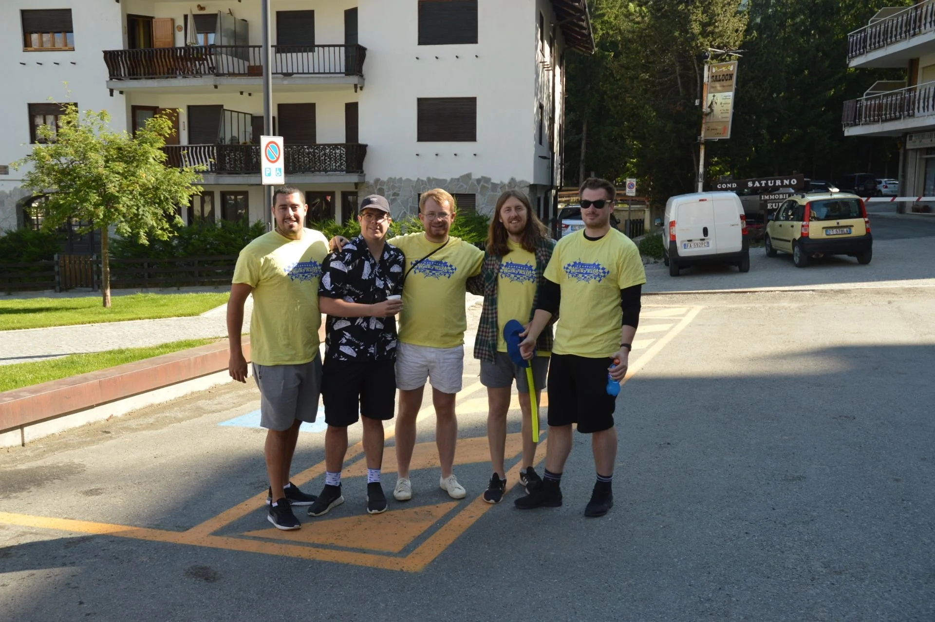 Group of five young men posing outdoors in a parking lot, with some of them wearing matching yellow t-shirts, and holding various items like water bottles and a pool noodle. Behind them are apartment buildings, parked cars, and trees.
