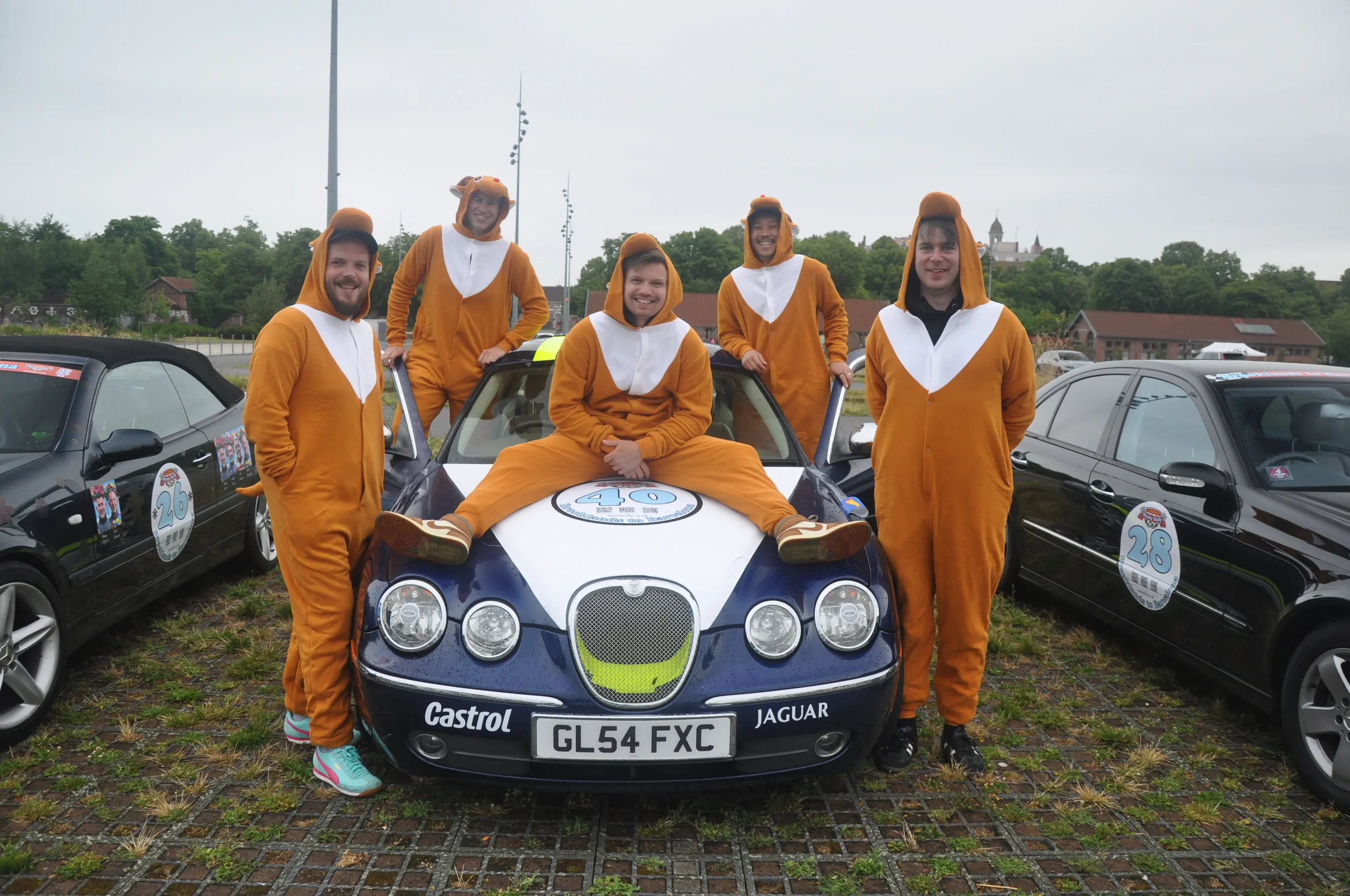 Group of six people dressed in lion and lioness costumes posing around a blue Jaguar race car with the number 40 on the hood, parked among other cars at a race event.