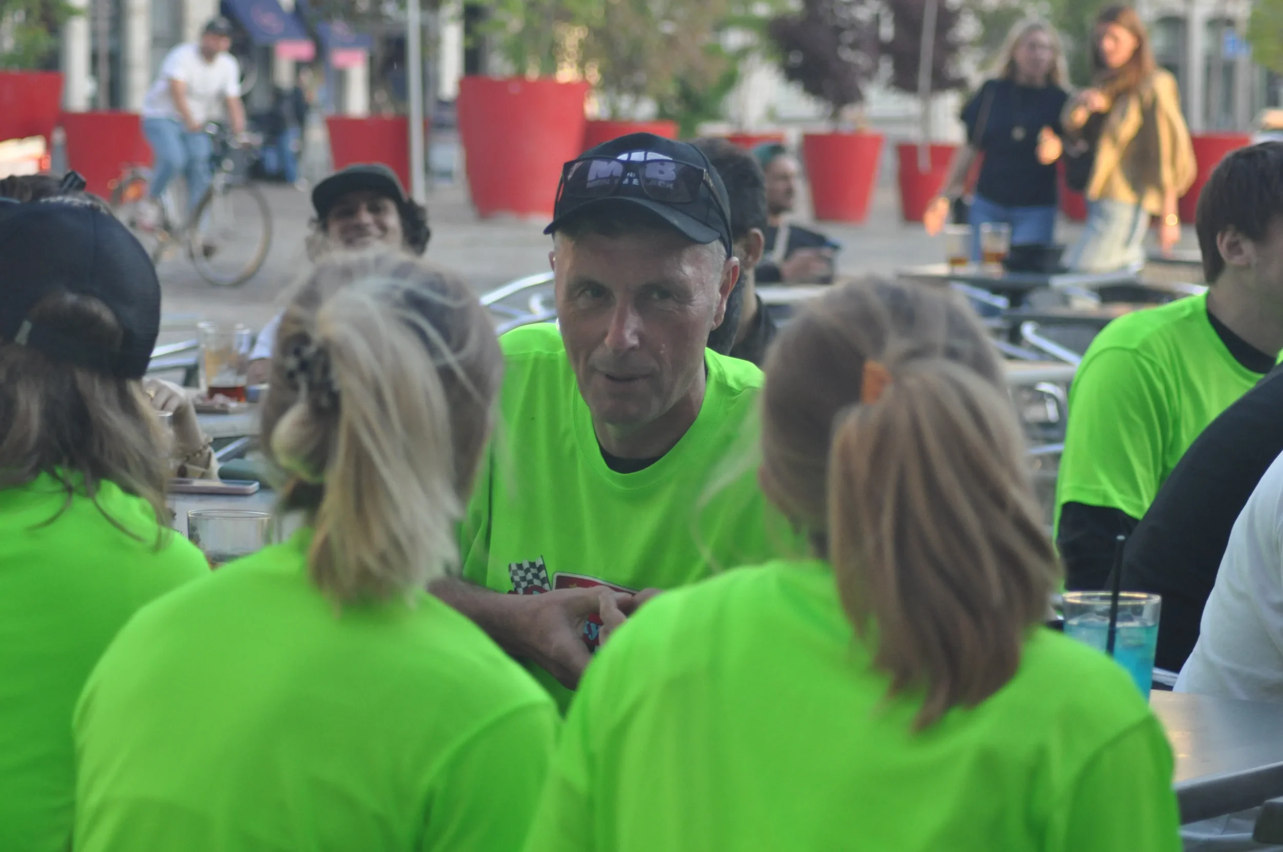 A man in a black cap and lime green shirt talking to a group of women also in lime green shirts at an outdoor seating area.