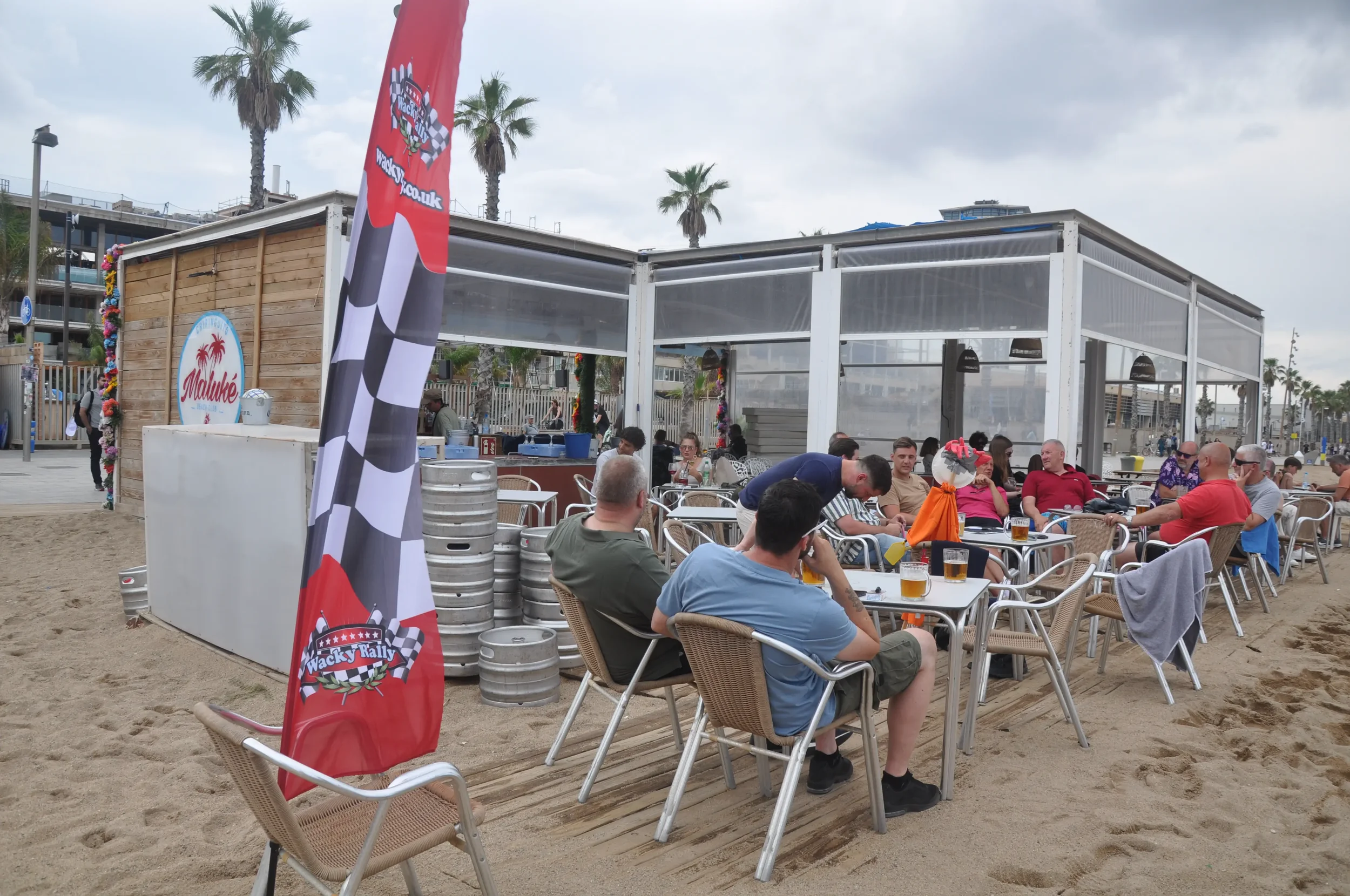 Group of people sitting at tables with drinks at a beachside restaurant, with a surfing-themed flag and beer kegs outside.