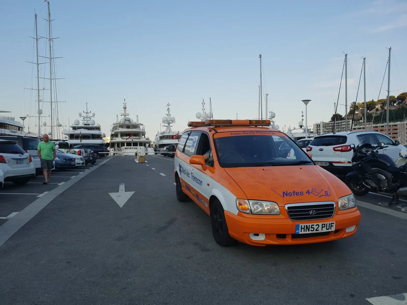 View of a marina with yachts in the background, a man walking on the parking lot, and an orange emergency vehicle with writing on it parked to the right.
