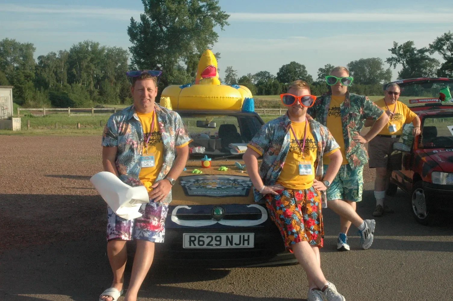 Four men dressed in beachwear, standing in front of a police car with a large inflatable duck on top. The men are wearing Hawaiian shirts, yellow t-shirts, colorful shorts, and sunglasses, posing outdoors during a sunny day.