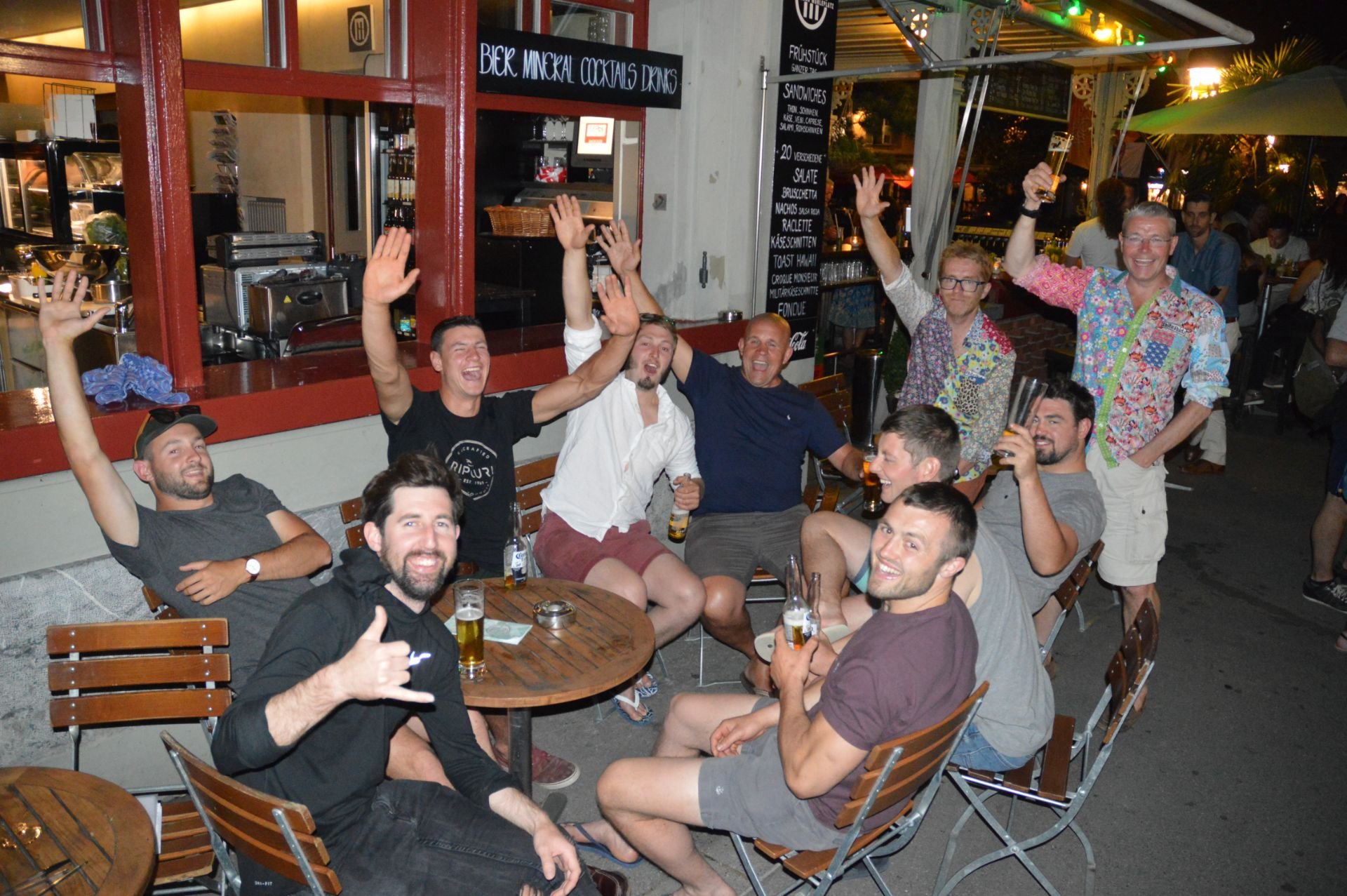Group of friends sitting at an outdoor bar or restaurant, smiling, some raising their hands or drinks, celebrating.