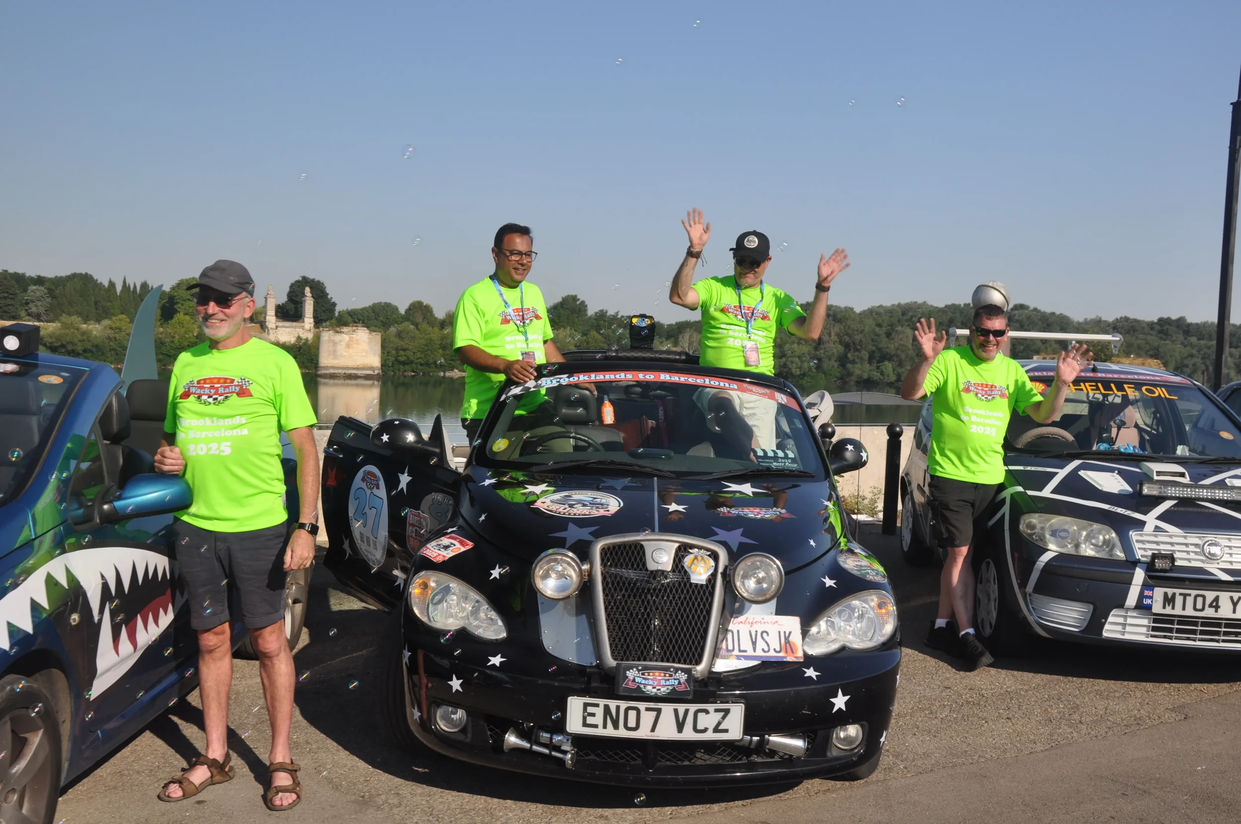 Four men in bright green t-shirts standing beside and on top of decorated rally cars, waving and smiling in a scenic outdoor location near a river or lake.