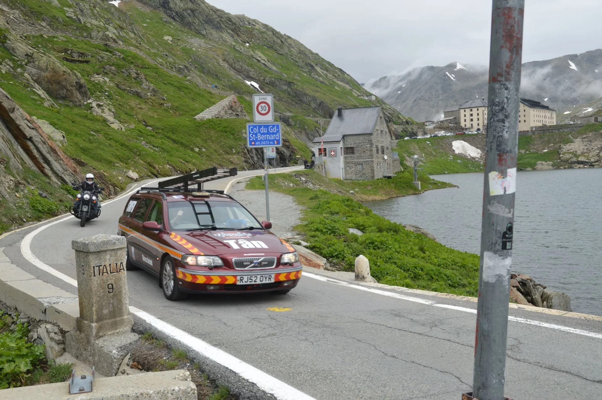 A scenic mountain road beside a lake with a stone building, road signs, a parked car, and a motorcycle with rider, in a foggy, mountainous landscape.