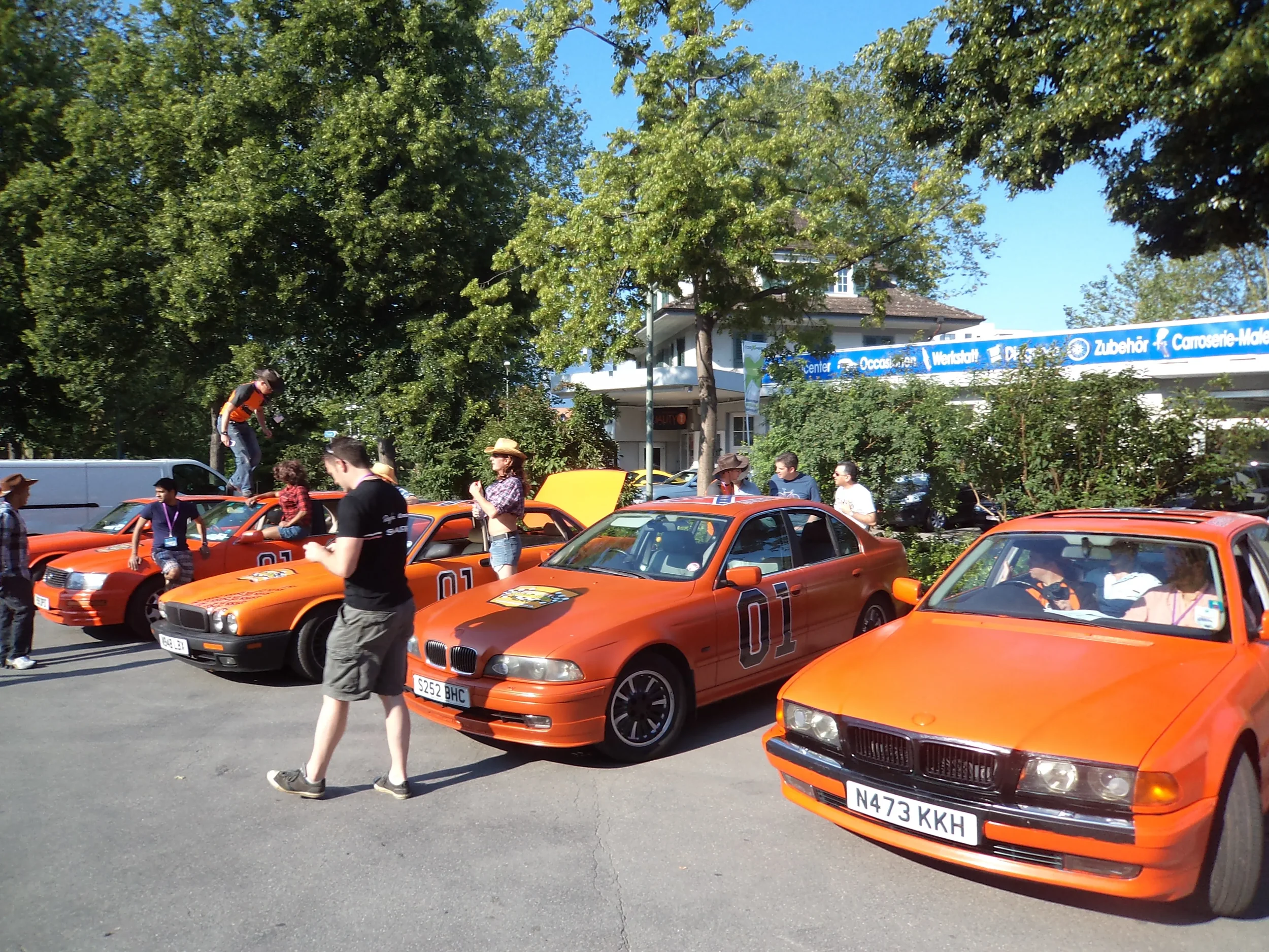 Multiple orange cars from the movie 'Cars,' with people walking and standing around, some taking pictures, on a sunny day with trees in the background.