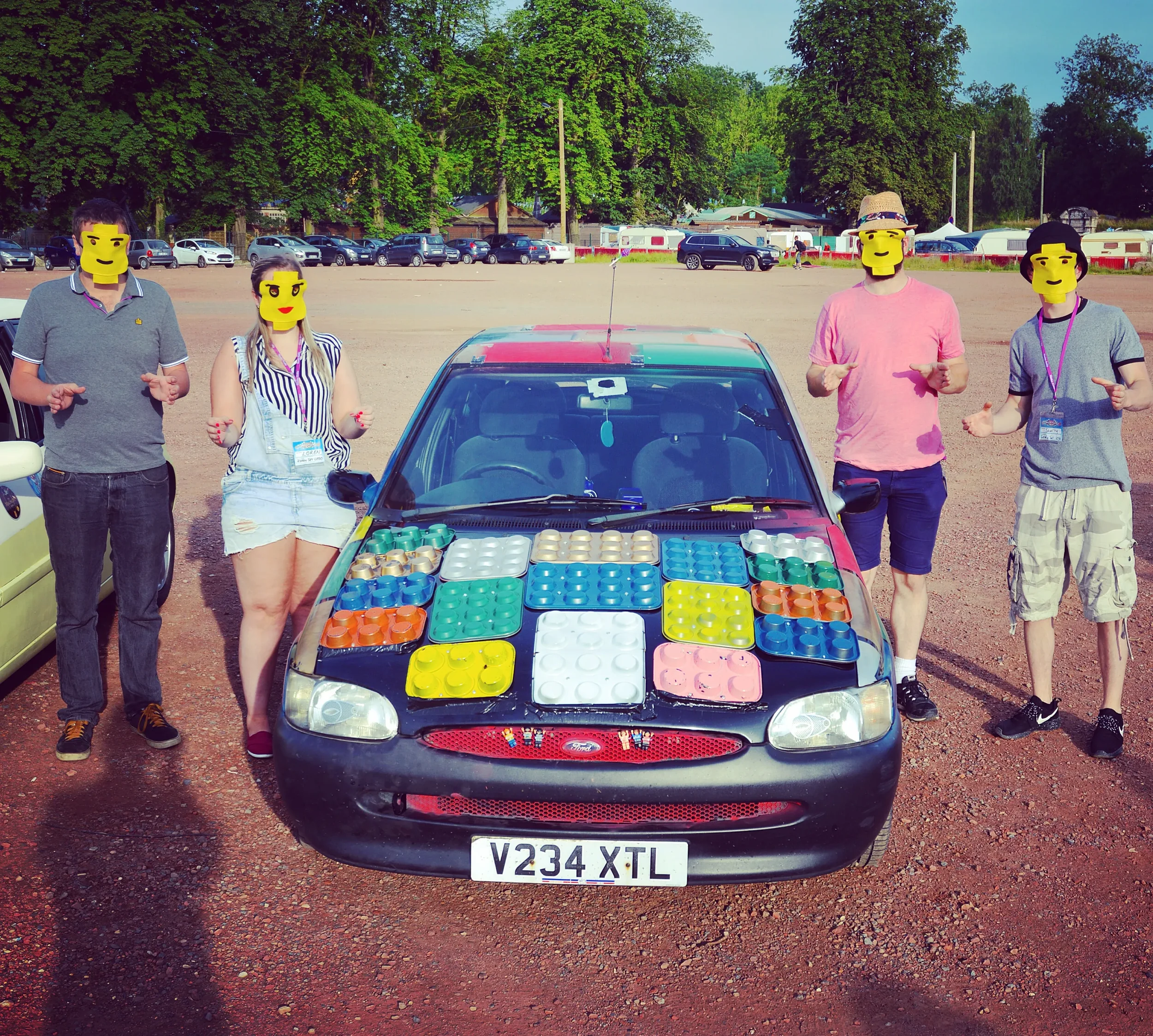 Group of five people standing around a black car with colorful Lego-style building blocks on the hood, in a parking lot with trees in the background, all wearing yellow emoji masks.