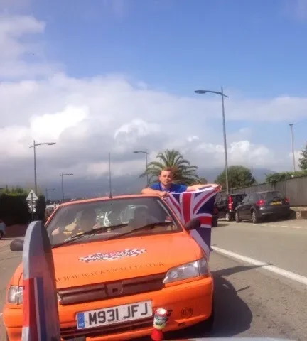 Person holding a United Kingdom flag standing next to an orange car on a street under a cloudy sky.