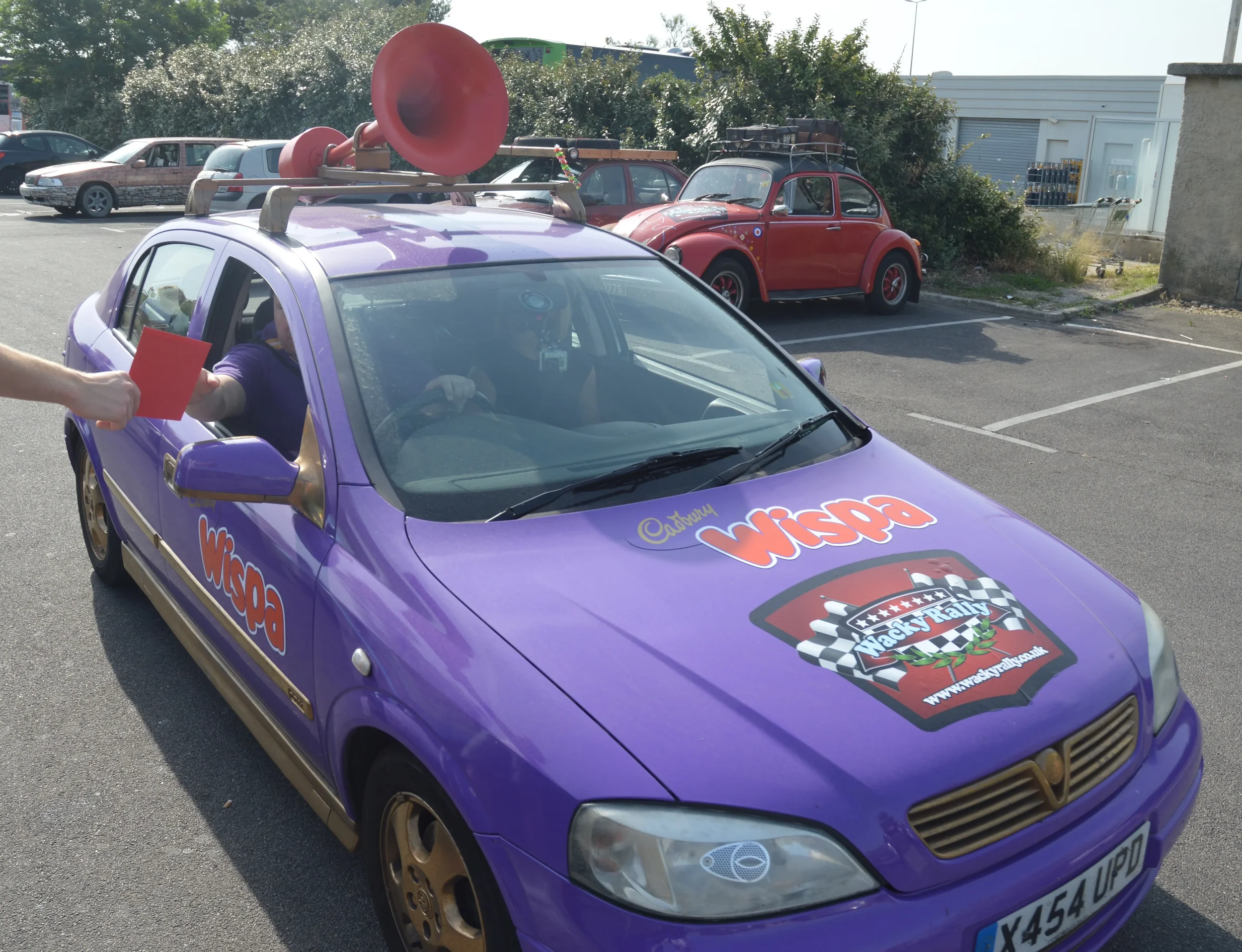 Purple race car with Red Bull horn on roof being handed a red card by a person. The car has 