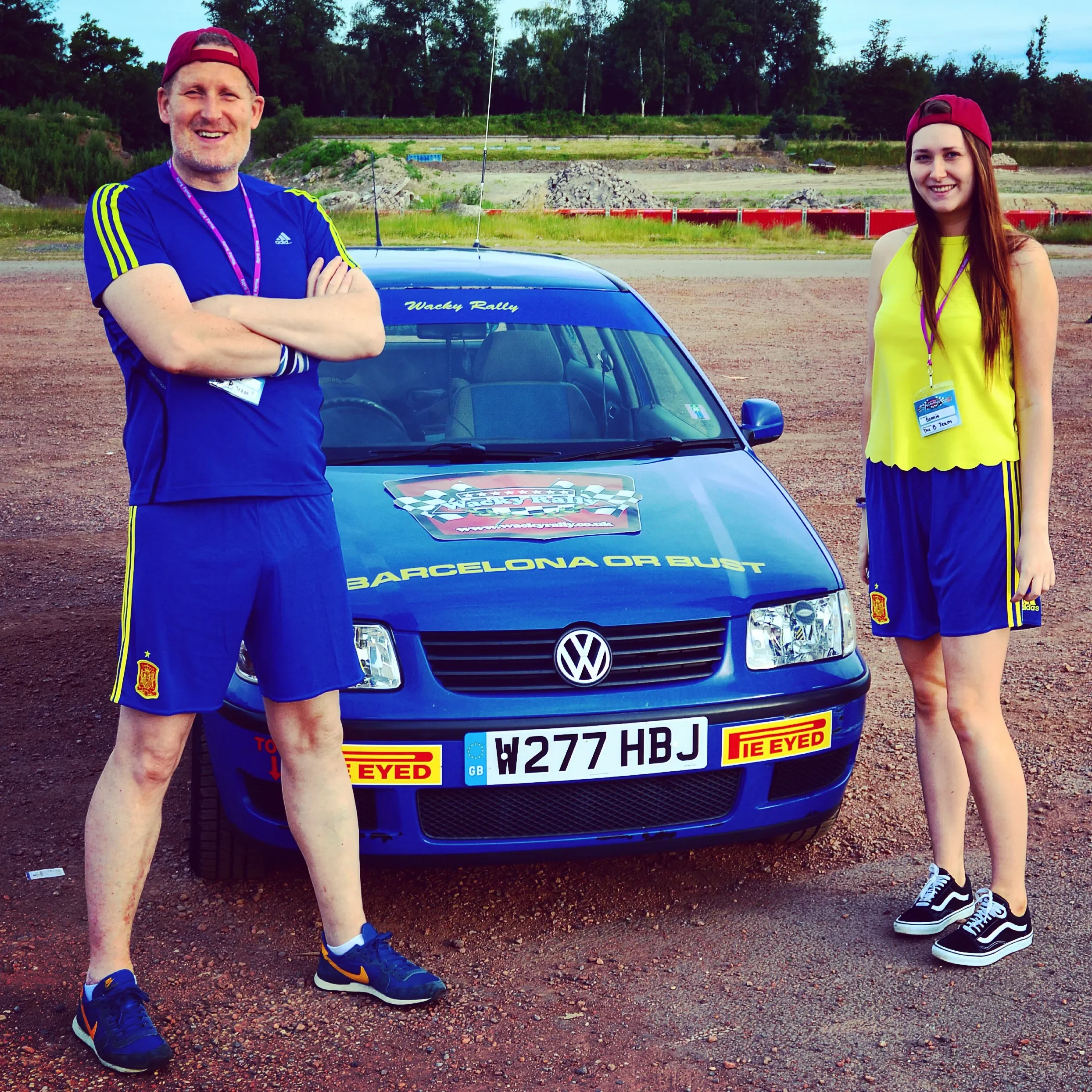 A man and a woman standing in front of a blue Volkswagen car on a dirt surface, both wearing sports attire and red caps, with a background of trees and an open field, appearing at a rally event.