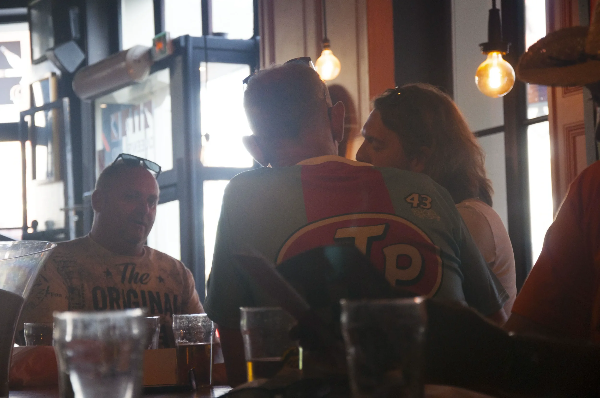 Two men are engaged in a conversation, seen from behind, in a dimly lit restaurant or bar. One man is wearing sunglasses on his head and a shirt with the number 43, while the other man has long hair. There are glasses and bottles on the table, and wa