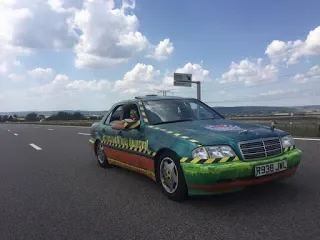A car with a green and yellow flame paint job driving on a highway under a partly cloudy sky.