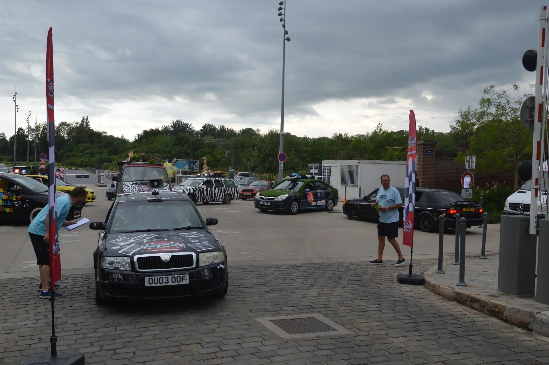 Festival parking lot with decorated cars, banners, and people, some examining cars and others walking, under a cloudy sky.
