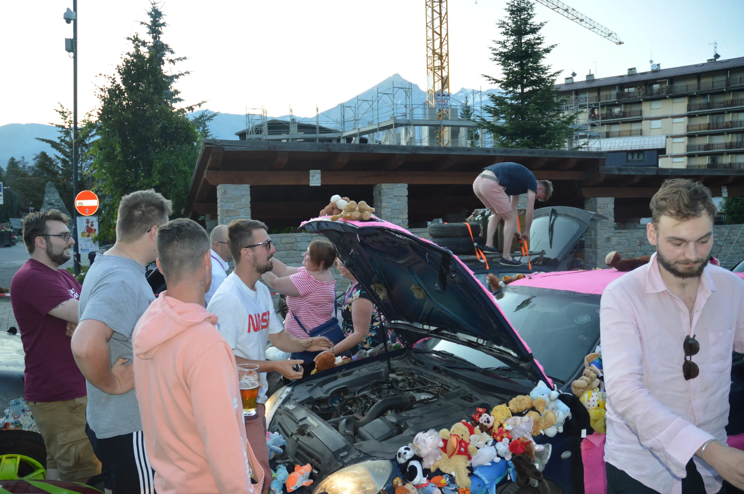 Group of people gathering around decorated cars with stuffed animals and pink accents in a parking lot outdoors, mountains and trees in the background.
