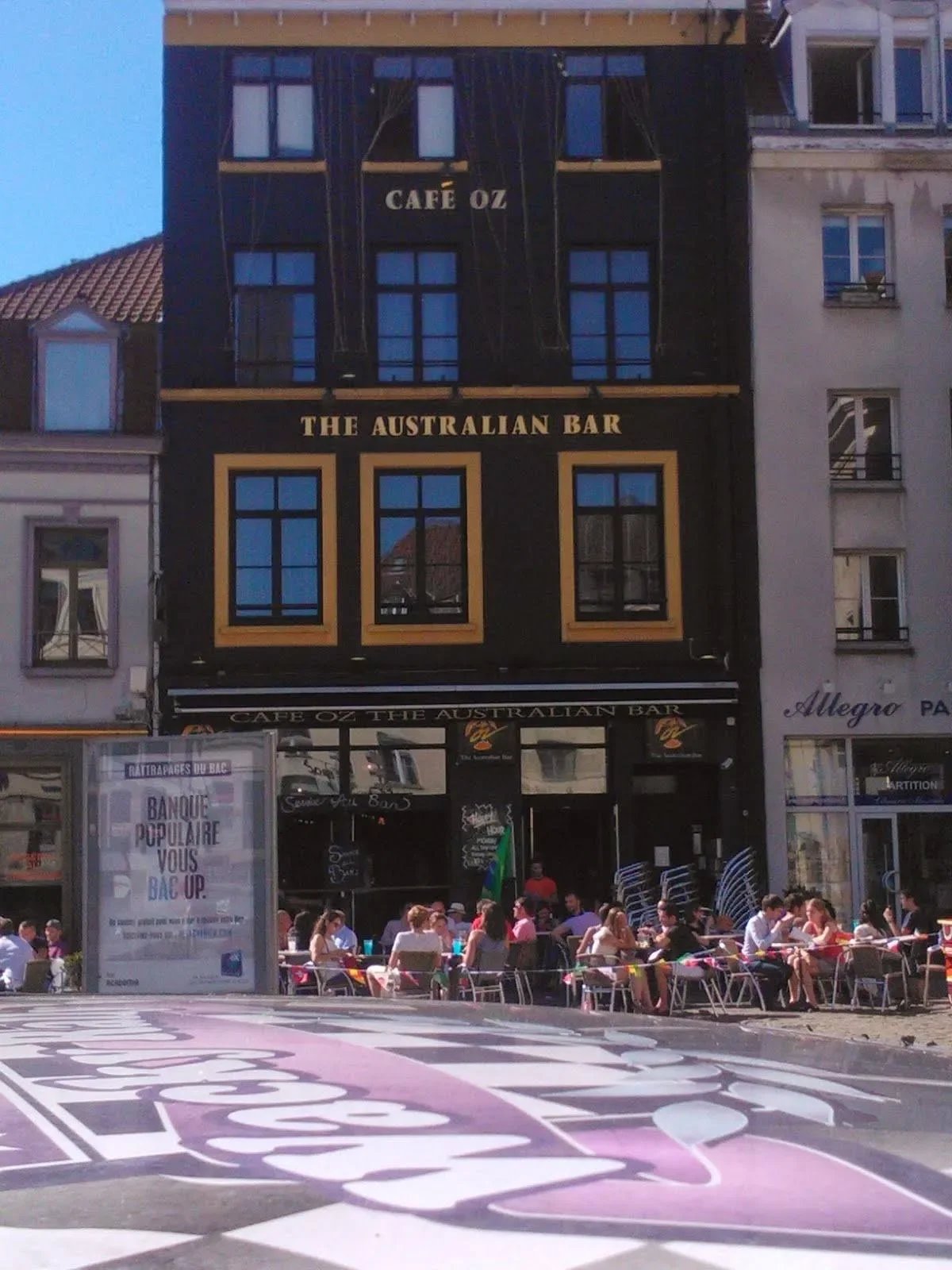Exterior view of Cafe Oz at The Australian Bar, showing a black building with gold accents and multiple windows, with outdoor seating filled with people dining on a sunny day.