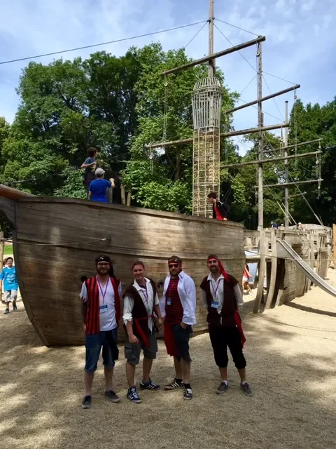 Four men standing in front of a large wooden pirate ship playground with children climbing on it and trees in the background.