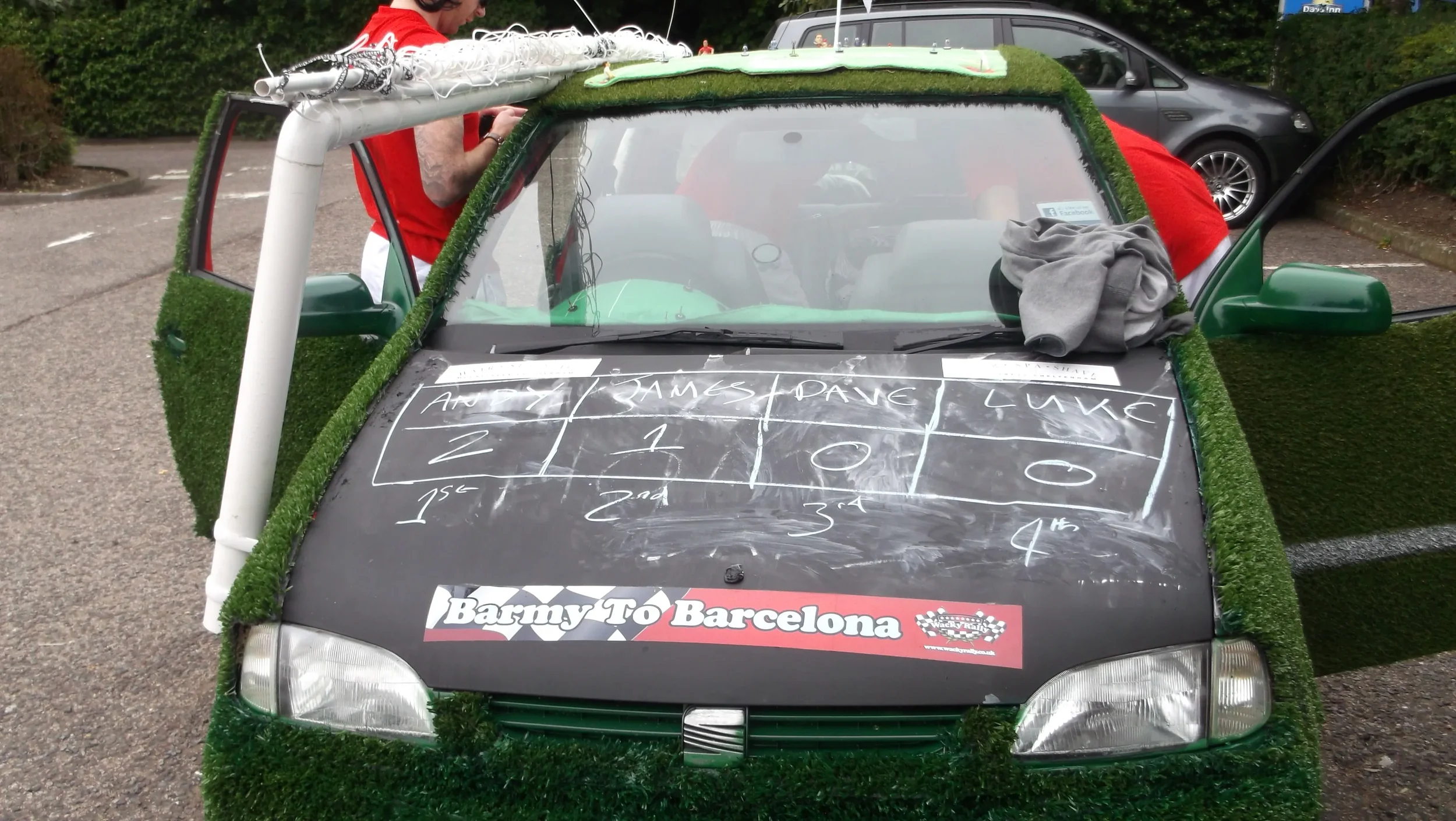 A car decorated with artificial grass for a racing event, featuring a chalkboard with betting odds and drivers' names, parked in a lot with people preparing it.