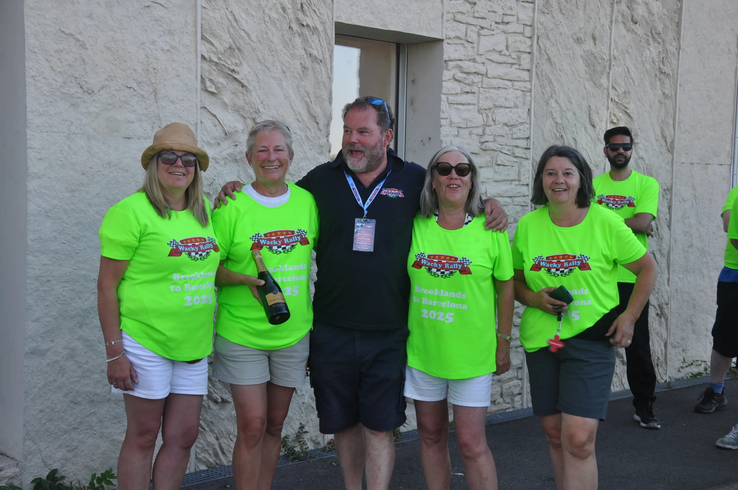 Group of people wearing bright green t-shirts with Wacky Rally logo, celebrating at an outdoor event, with one holding a bottle of champagne, smiling and posing for the photo.