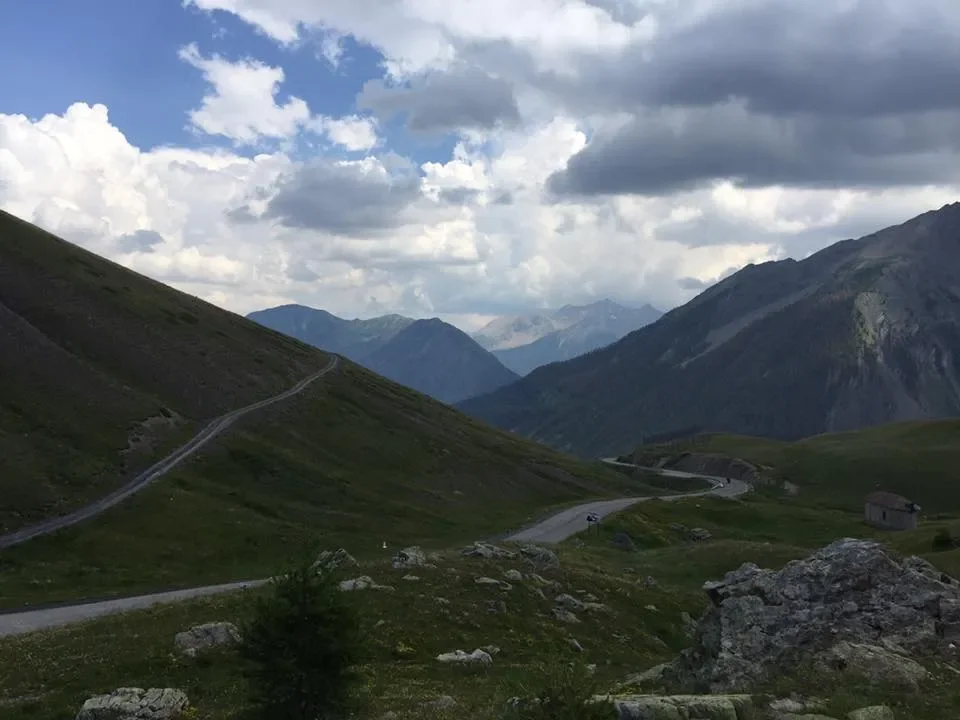 Mountain landscape with winding road, green grassy slopes, and cloudy sky.