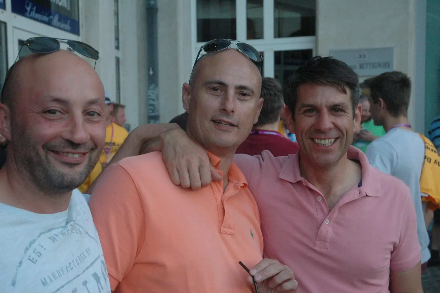 Three men smiling, standing outdoors among a crowd, with one man in an orange shirt and sunglasses on his head, and the others in pink and white shirts.
