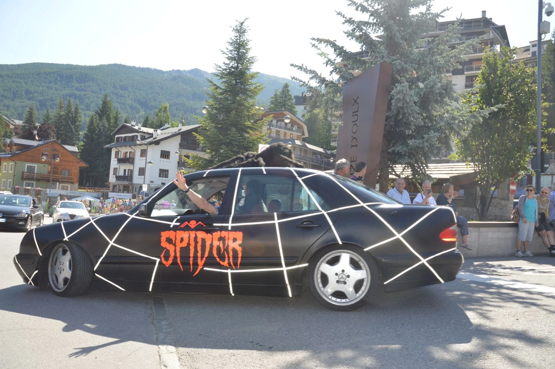 A black car with white web-like stripes and a red Spider-Man logo, with a person inside waving, parked on a city street with people and buildings in the background.