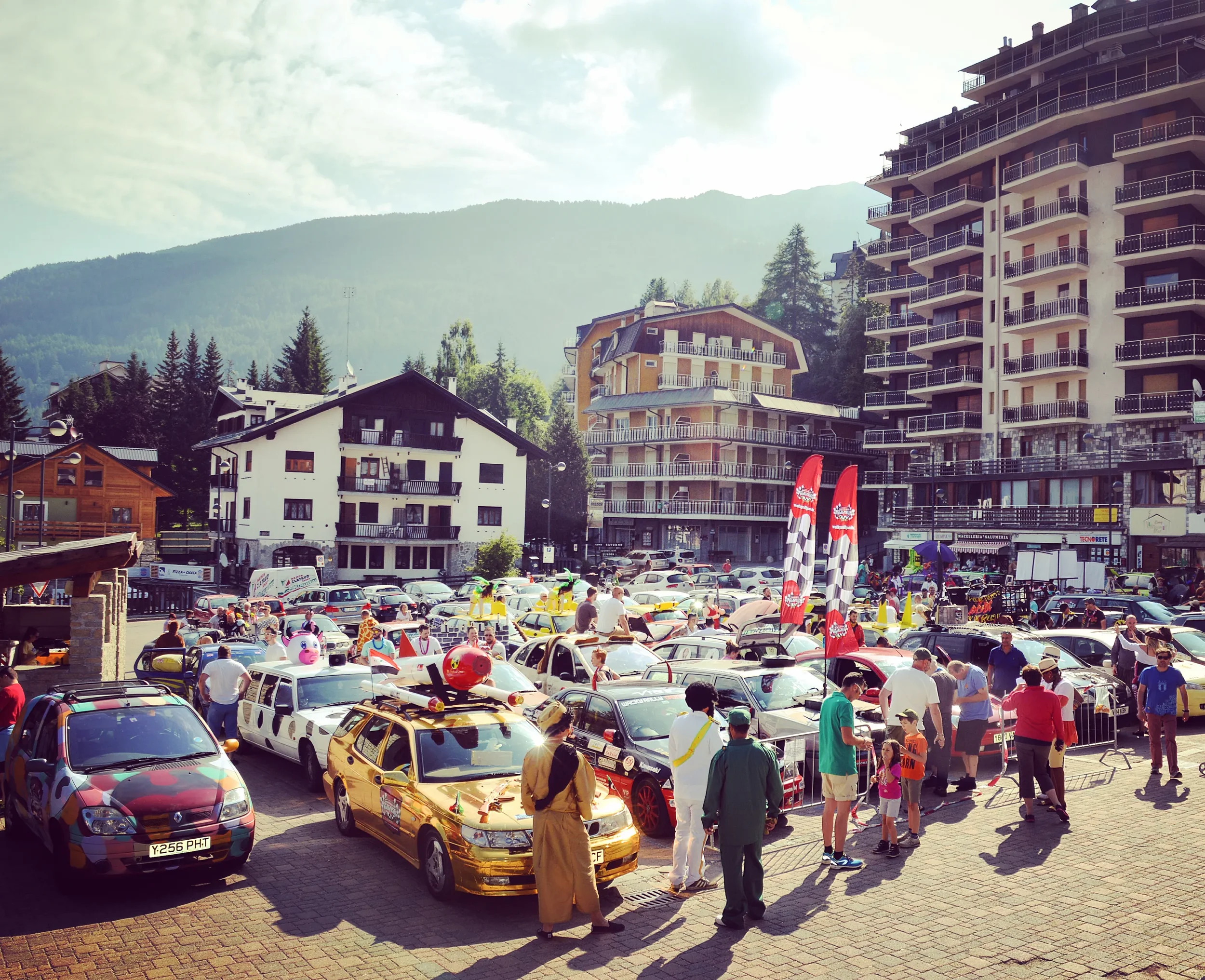 A lively outdoor event in a town square with decorated cars and spectators, surrounded by multi-story buildings and hills in the background.