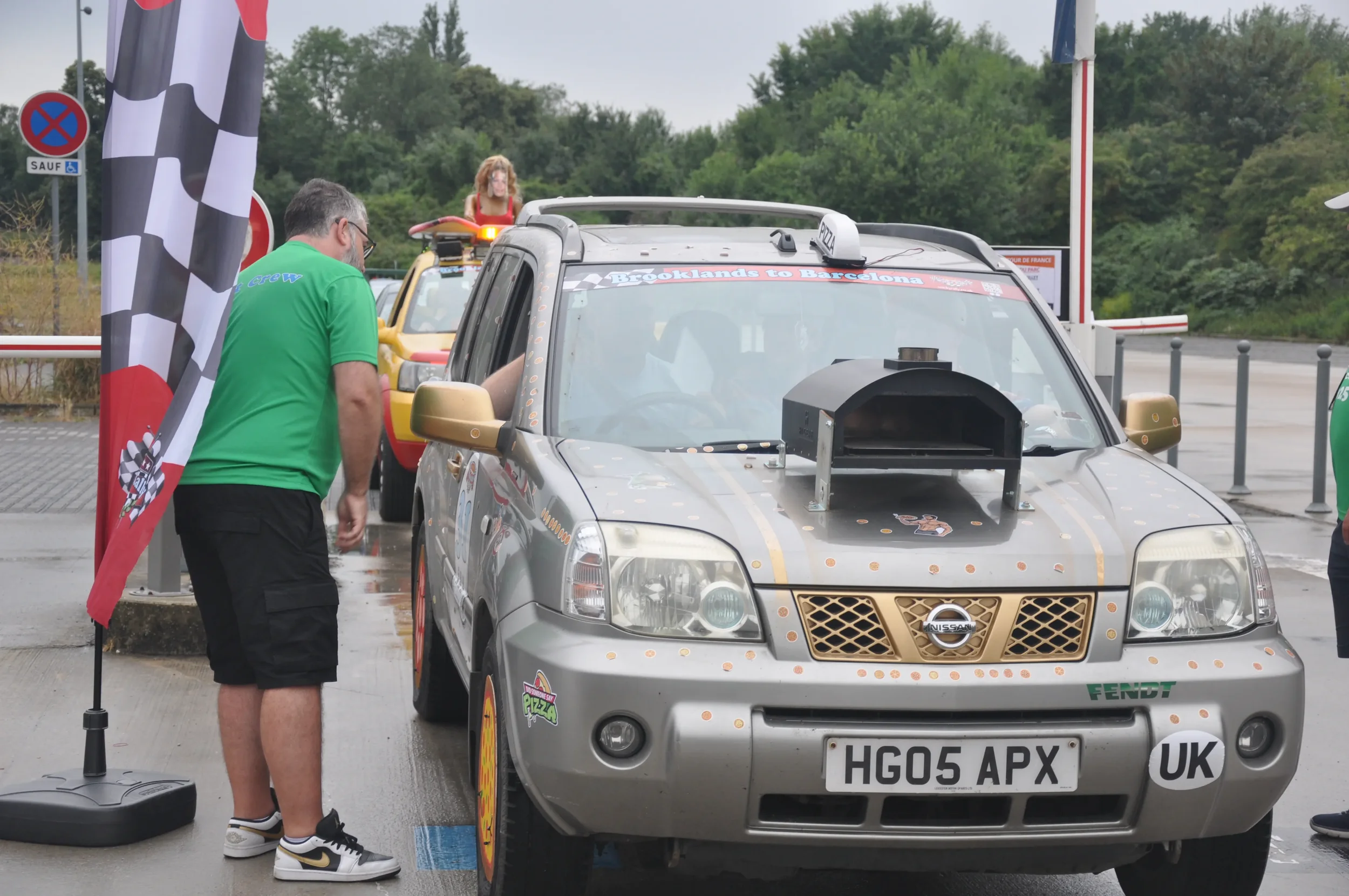 A silver Nissan SUV decorated with racing stickers, parked at a race event, with a man in a green shirt inspecting it and a checkered flag in the foreground.