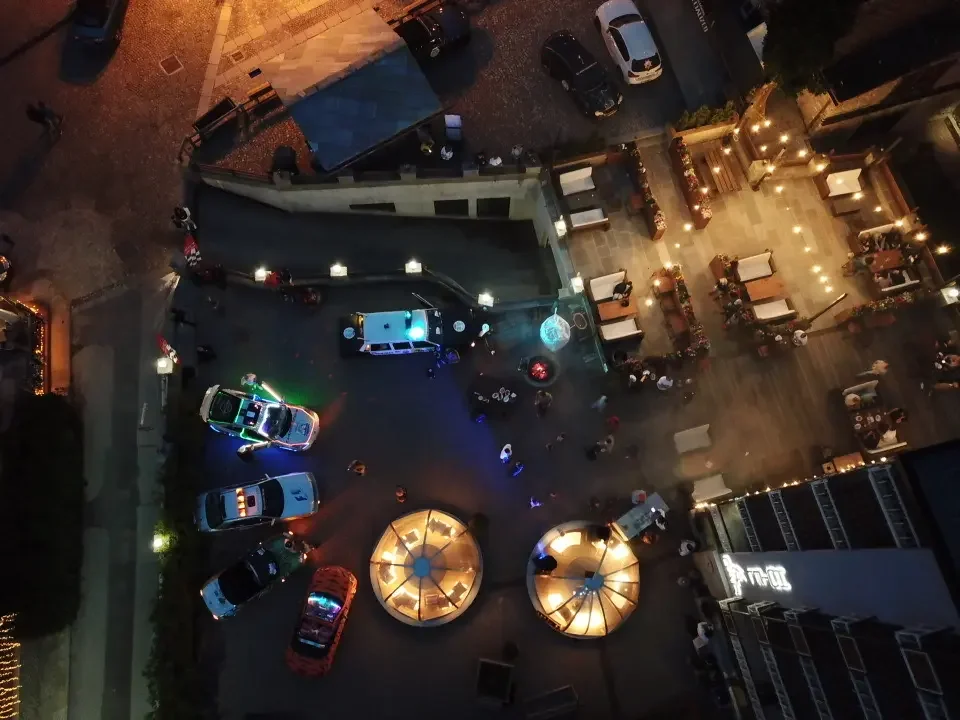 Night aerial view of a city street scene with police cars, a lit outdoor seating area with umbrellas, and people walking around.