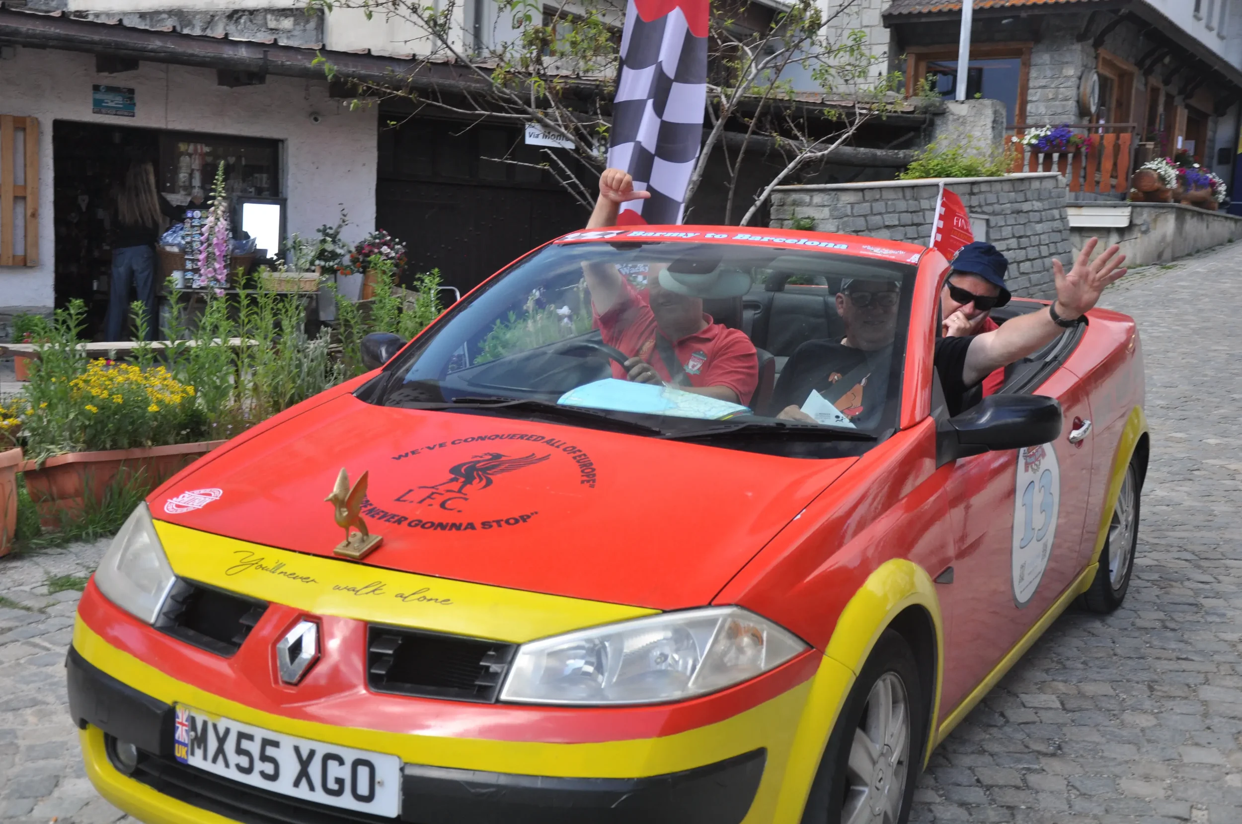 A red and yellow convertible Renault car with the number 13 on the side, driving on a cobblestone street, with two men inside waving and smiling. The car has various decals and a small golden angel statue on the hood.