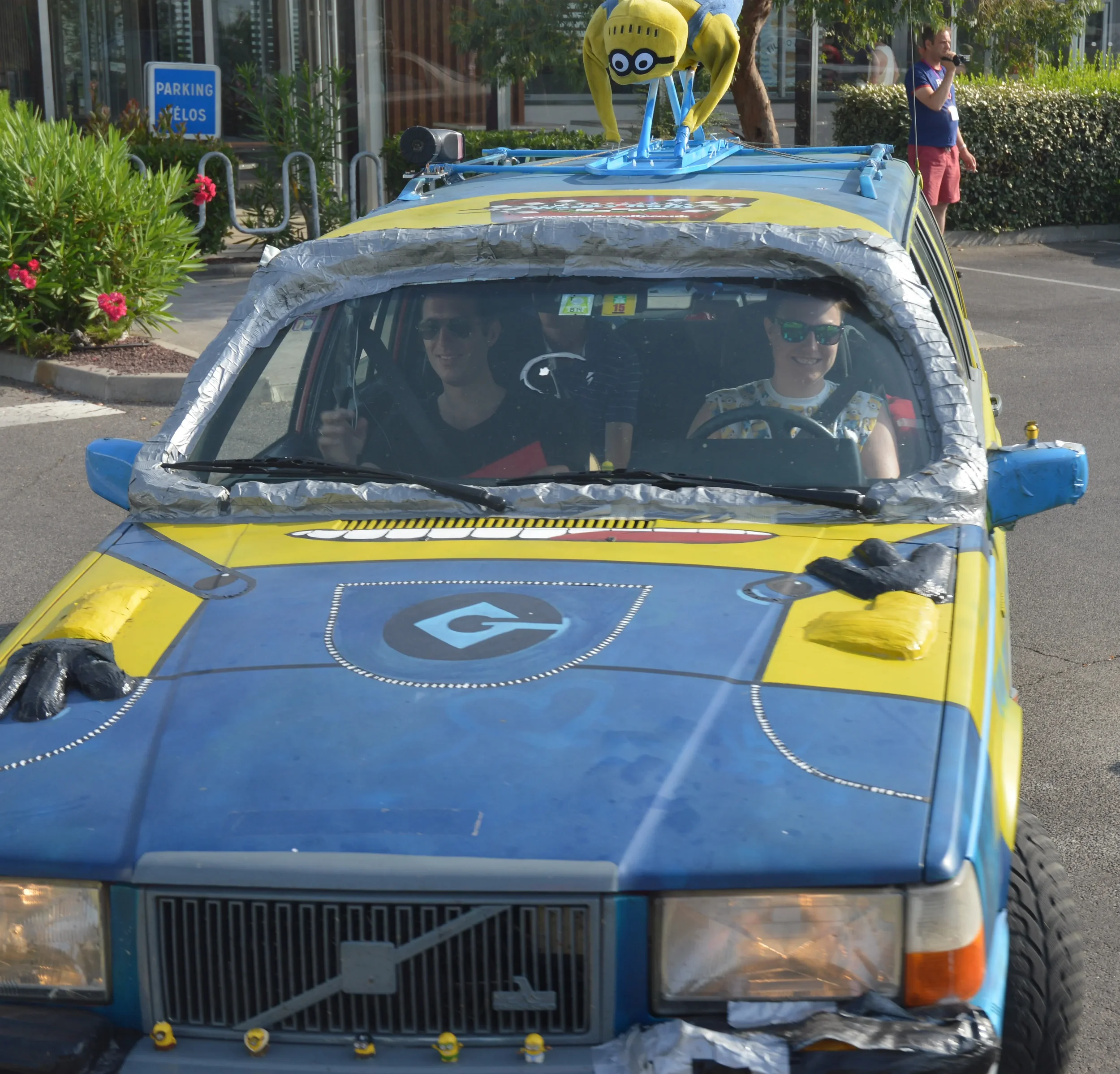 A yellow and blue themed car with a Minion figure on the roof, driven by two smiling people wearing sunglasses, parked in a lot with a person speaking into a microphone in the background.