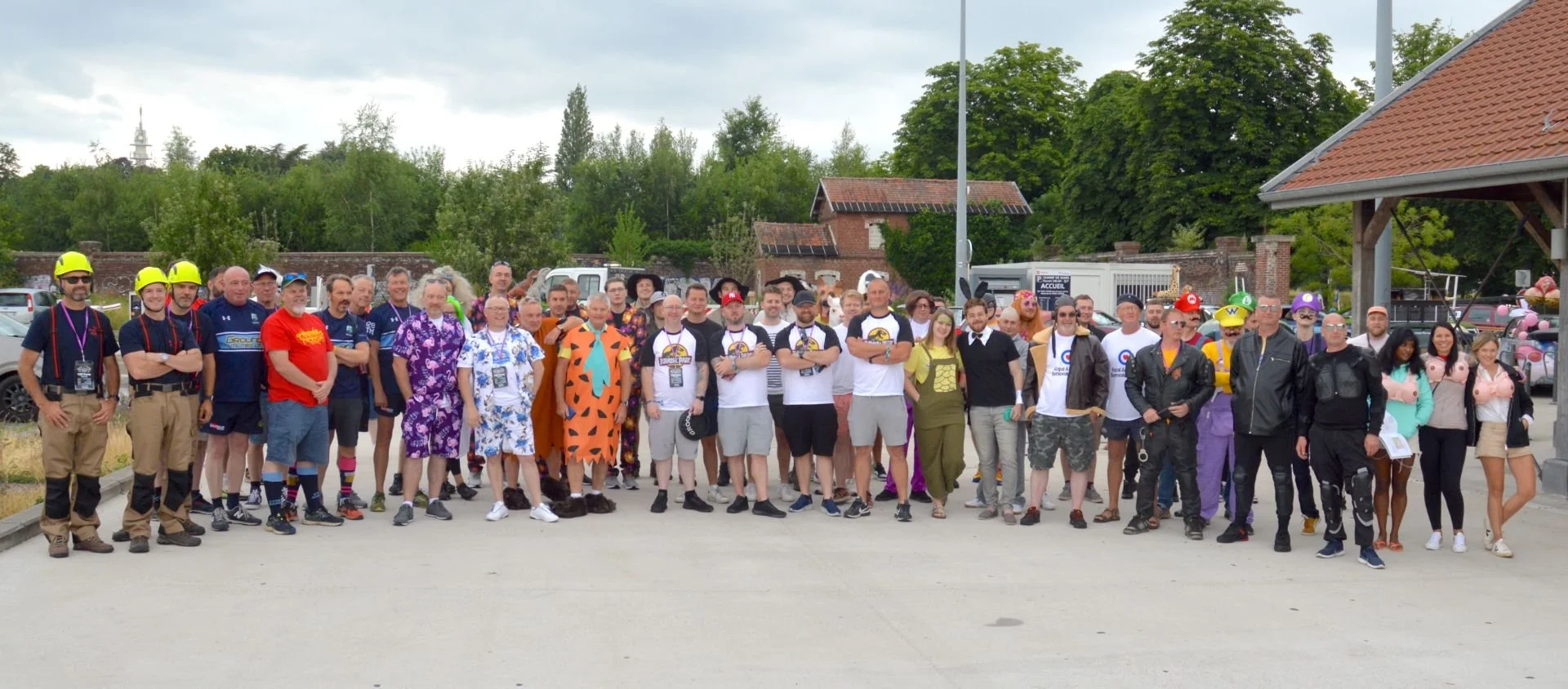 Group of people gathered outdoors, some dressed in costumes and others in casual wear, with a few wearing helmets, in front of trees and buildings.