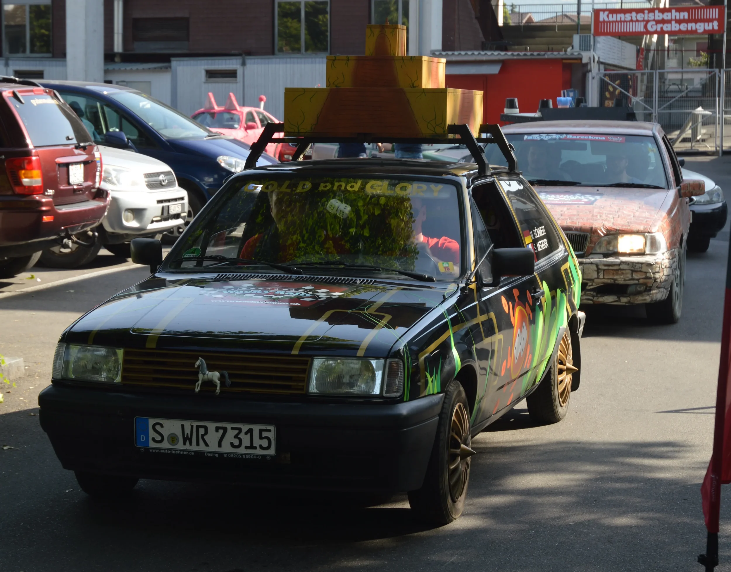 Colorful, graffiti-style decorated car with a horse figurine on its front grill, driving through a parking area with other cars and a red sign in the background.