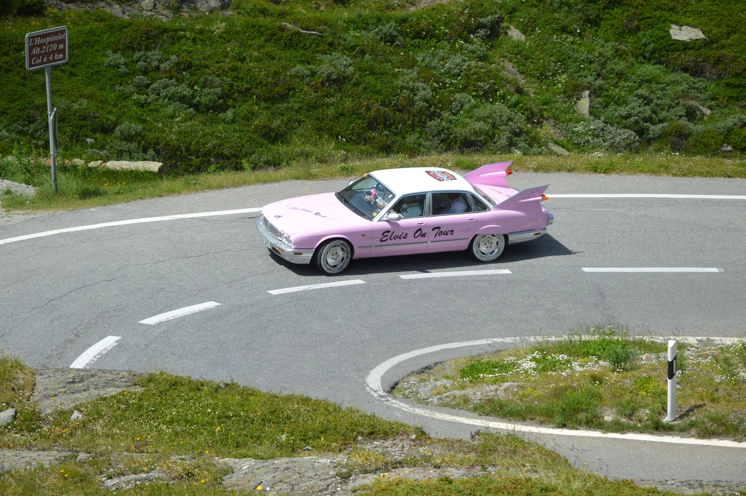 A pink and white car with 'Elvis On Tour' written on the side, driving along a winding mountain road with greenery and rocks in the background.
