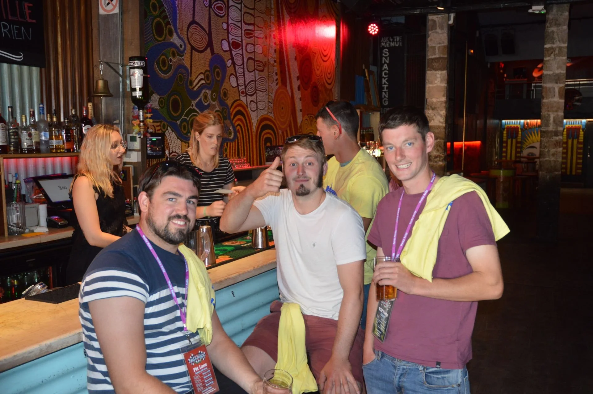 A group of five young men inside a bar, smiling and holding drinks, with a bartenders and colorful abstract wall art in the background.