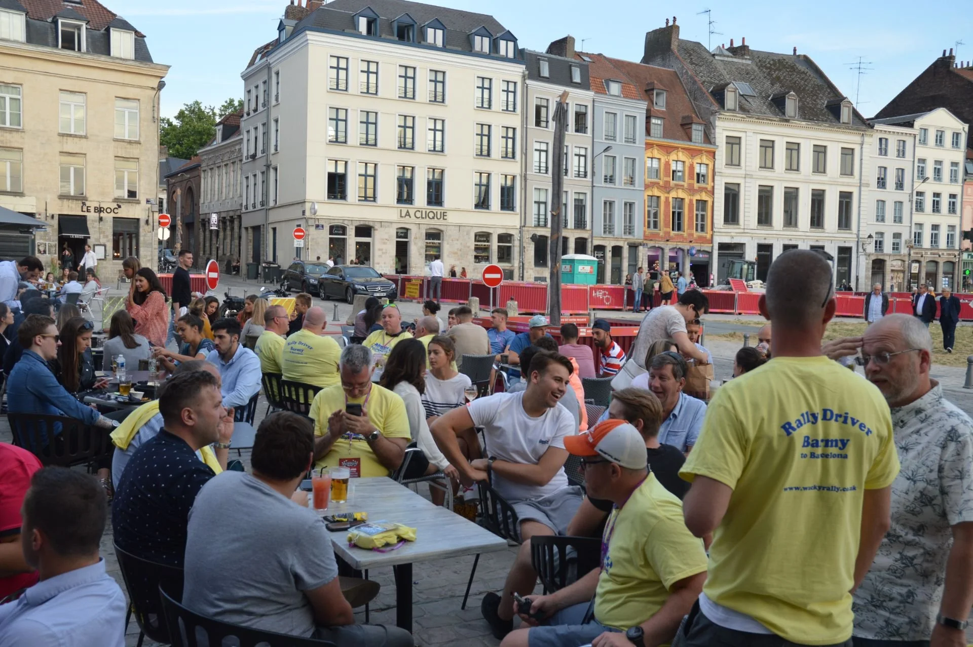 People gather at an outdoor café or event space in front of historic European-style buildings, some of which are under construction or renovation.