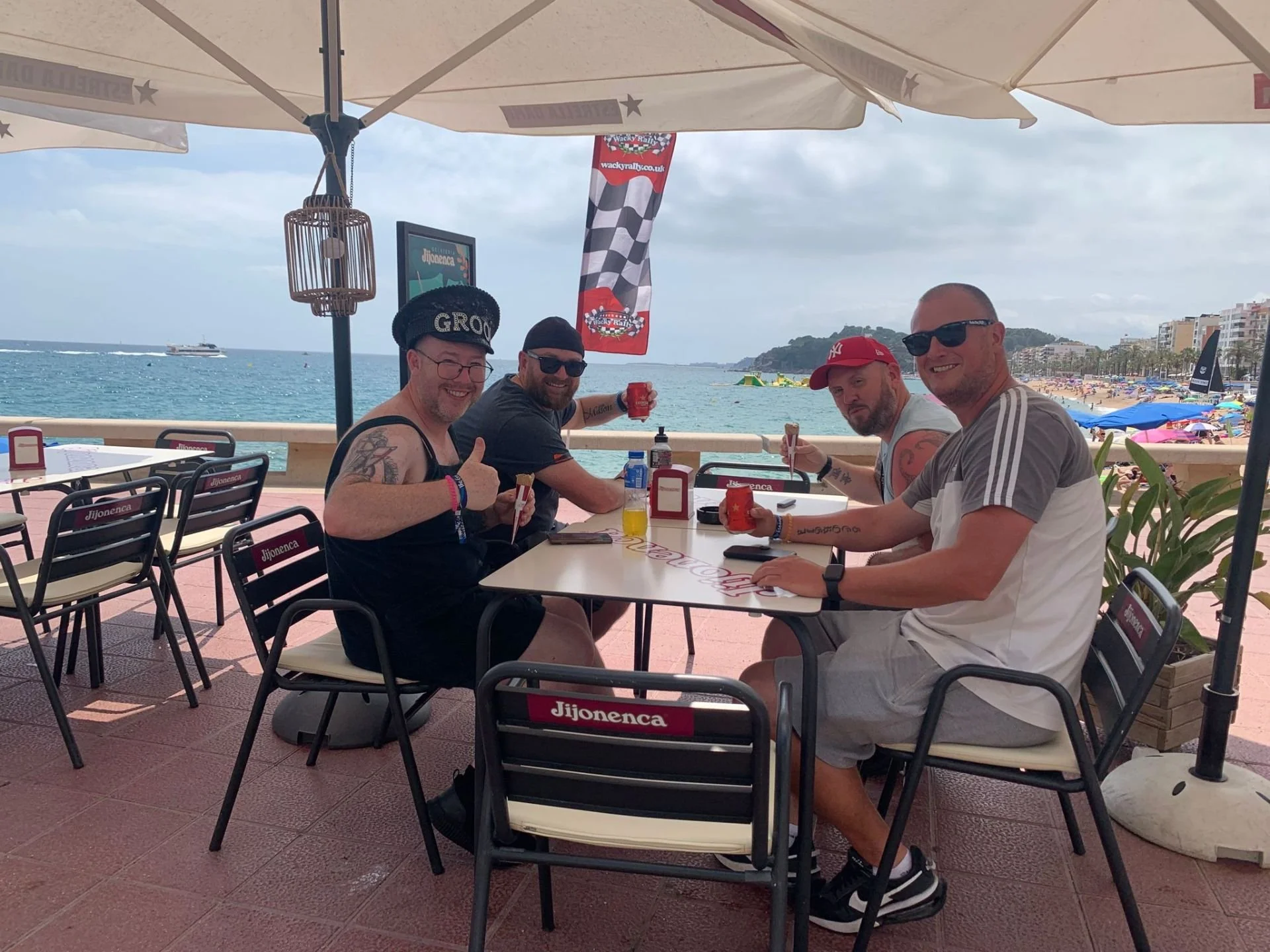 Four men sitting at a table with drinks and food under a large umbrella on a beachside restaurant patio, with the ocean, beach, and buildings in the background.