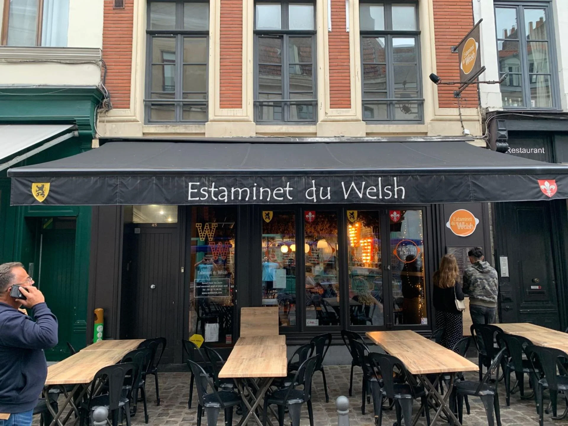 Front of a restaurant named 'Estaminet du Welsh' with outdoor seating and a couple of people standing near the entrance.