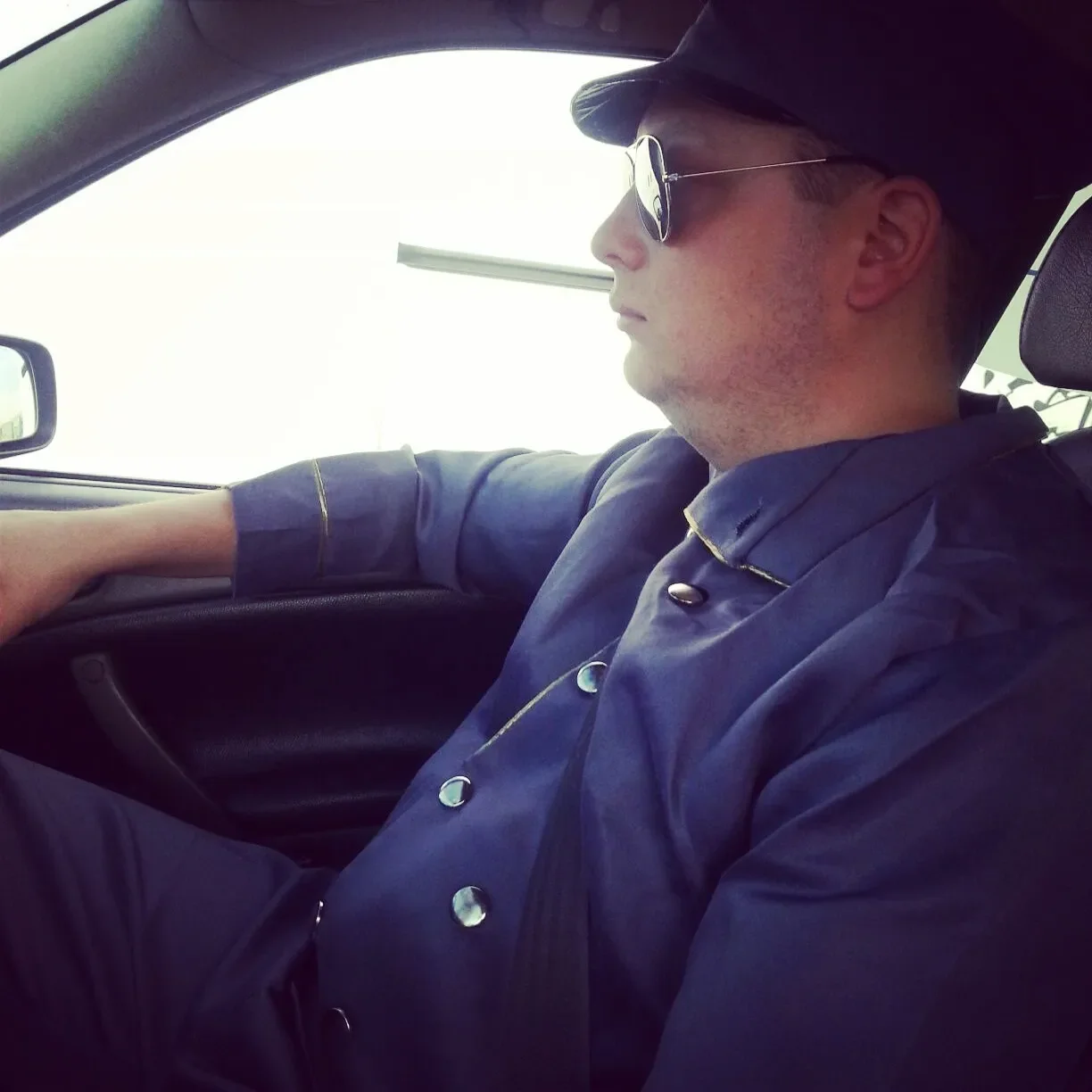 A man in sunglasses, a cowboy hat, and a dark shirt sitting in the driver's seat of a car, looking forward.