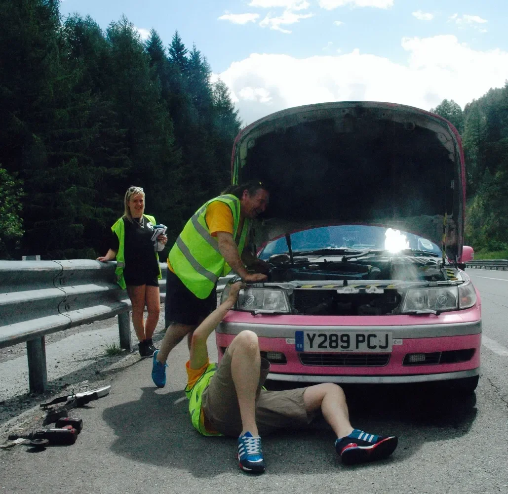 A car has broken down on the side of the road. Two men in safety vests are working under the open hood, while a woman with a notepad stands nearby. One man is sitting on the ground, working on the car, and the other is standing and inspecting the eng