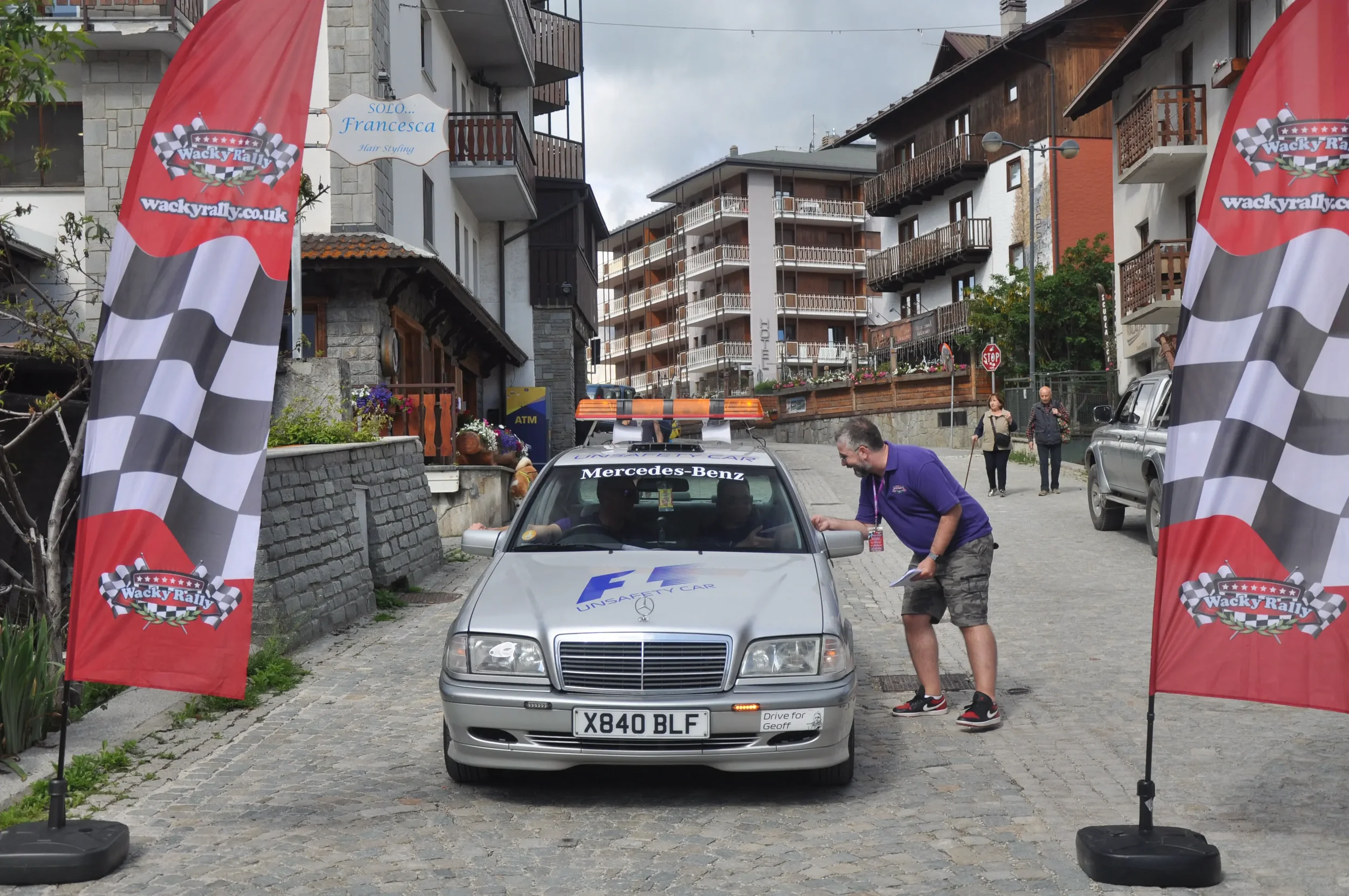 A silver Mercedes-Benz car parked under checkered flags with Wacky Rally logos at a race event, with a man talking to the driver and spectators in the background.