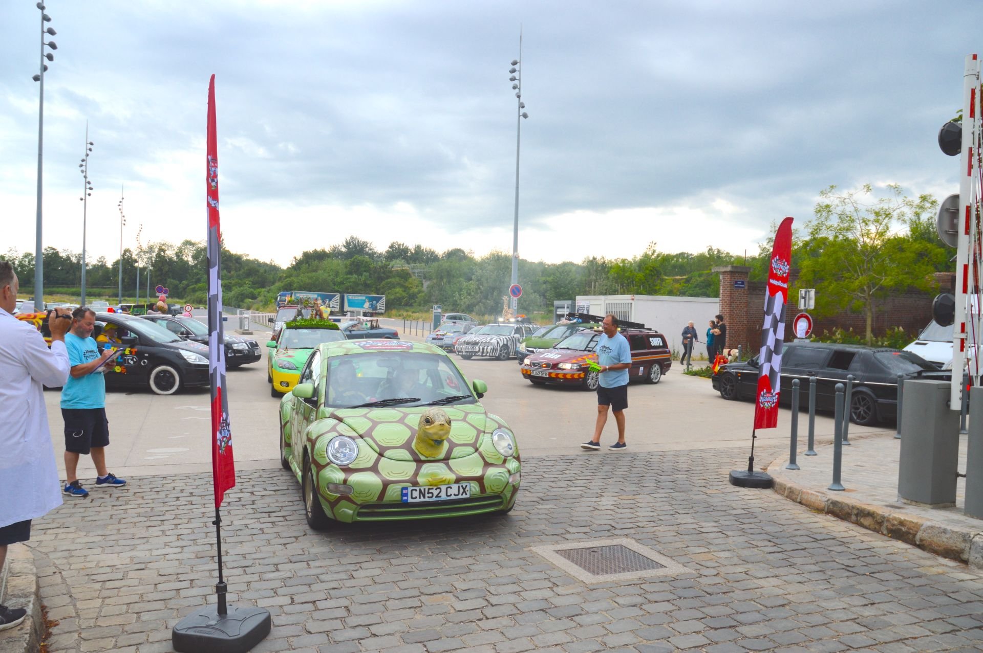 A green car decorated with a turtle shell pattern and a turtle face on the hood, parked in a lot with other cars and people taking photos. There are flags and people, some in casual and some in medical attire, with overcast skies in the background.